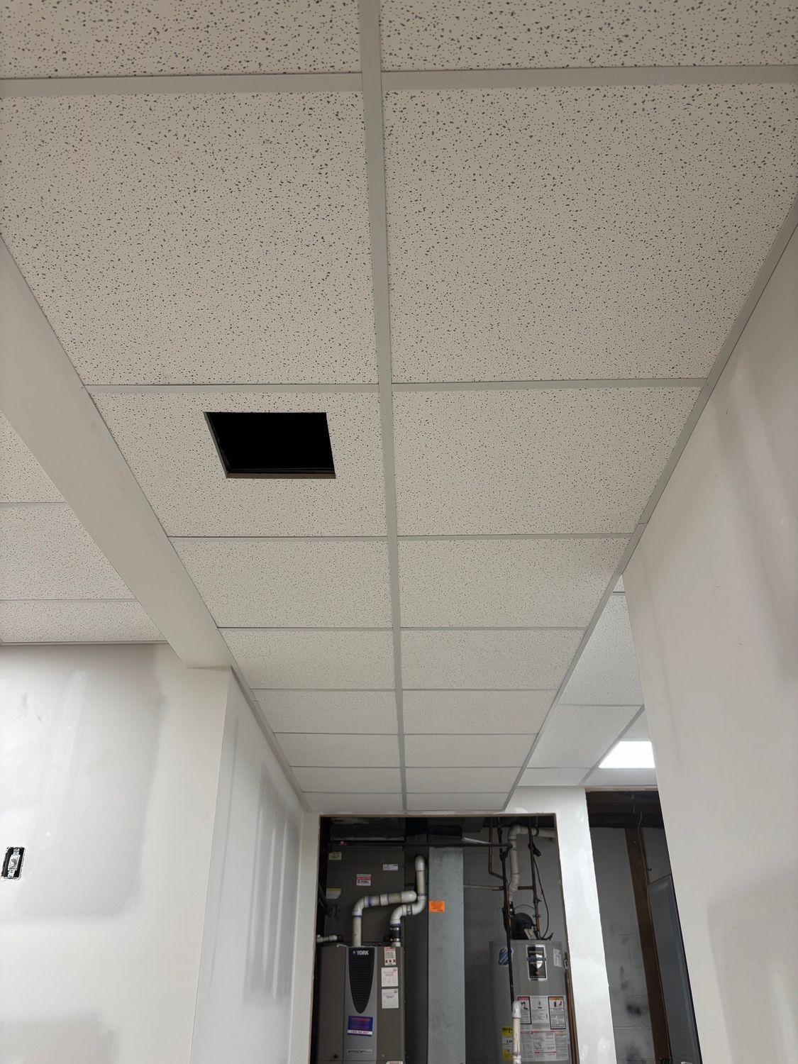 White drop ceiling with a missing tile, in a white-walled hallway with HVAC equipment visible in the distance.