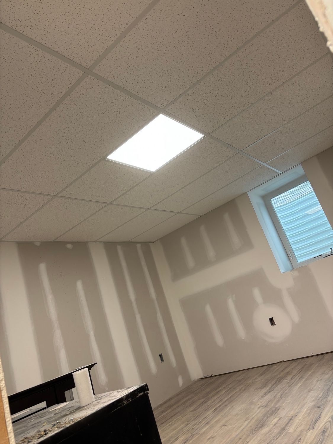 Room with unfinished walls, drop ceiling, window, and wood-look flooring; light fixture illuminated.