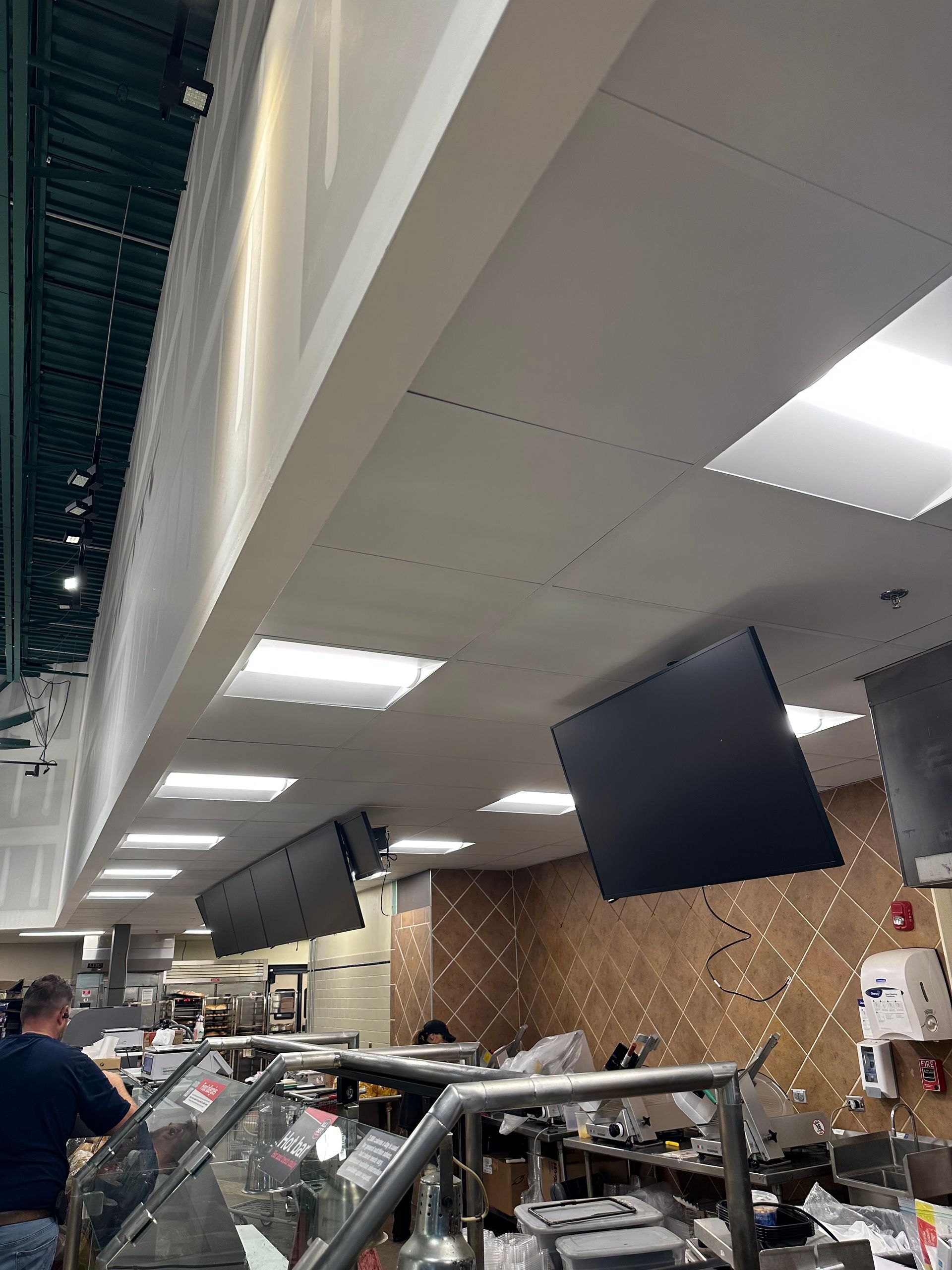 Ceiling with recessed lights, several hanging monitors, and a person near a food display case.