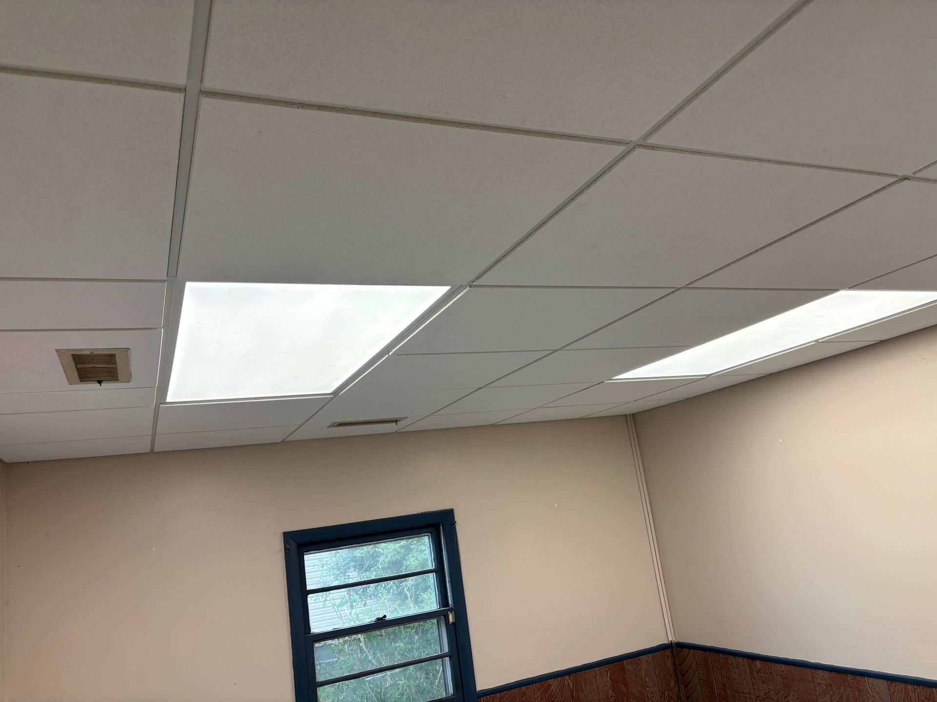A room with a drop ceiling, window, and beige walls. Overhead fluorescent lights illuminate the space.