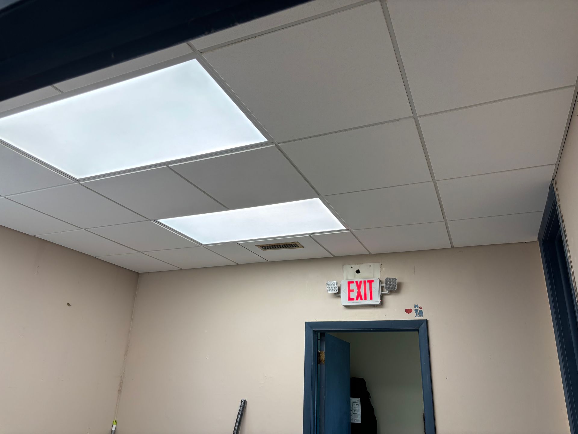 An office ceiling with fluorescent lights and an exit sign above a doorway. Beige walls.