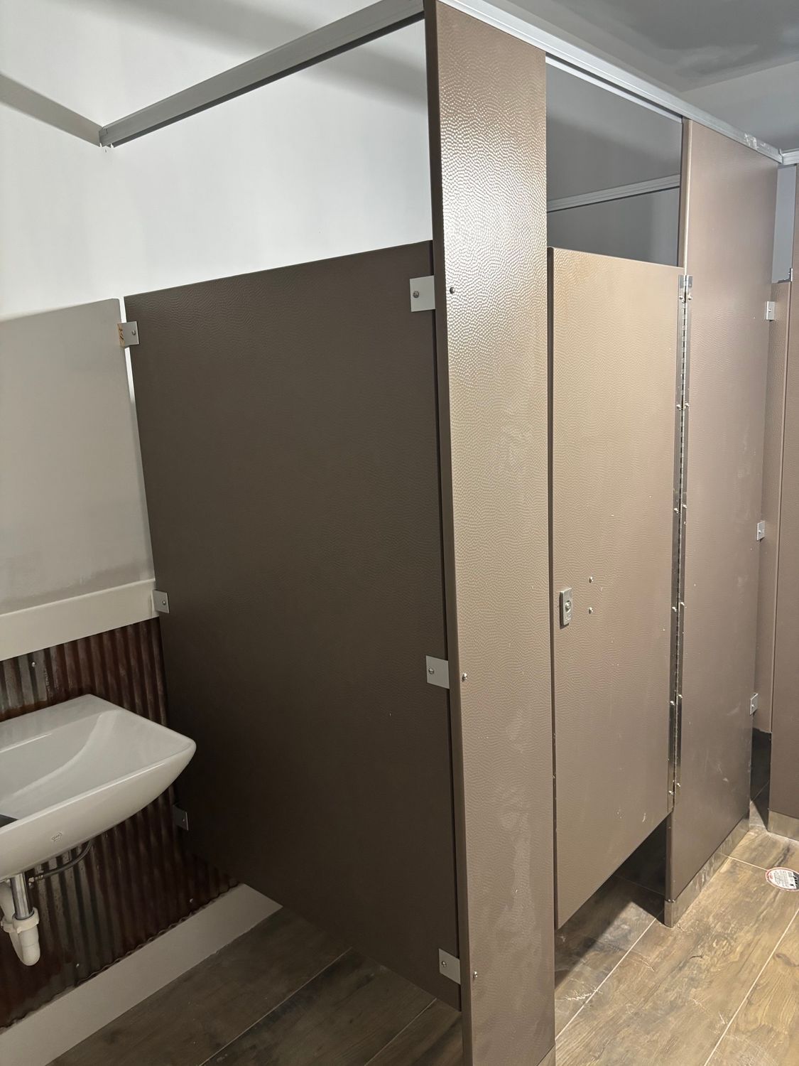 Bathroom stalls in a public restroom. Brown doors, white sink, and light floor.