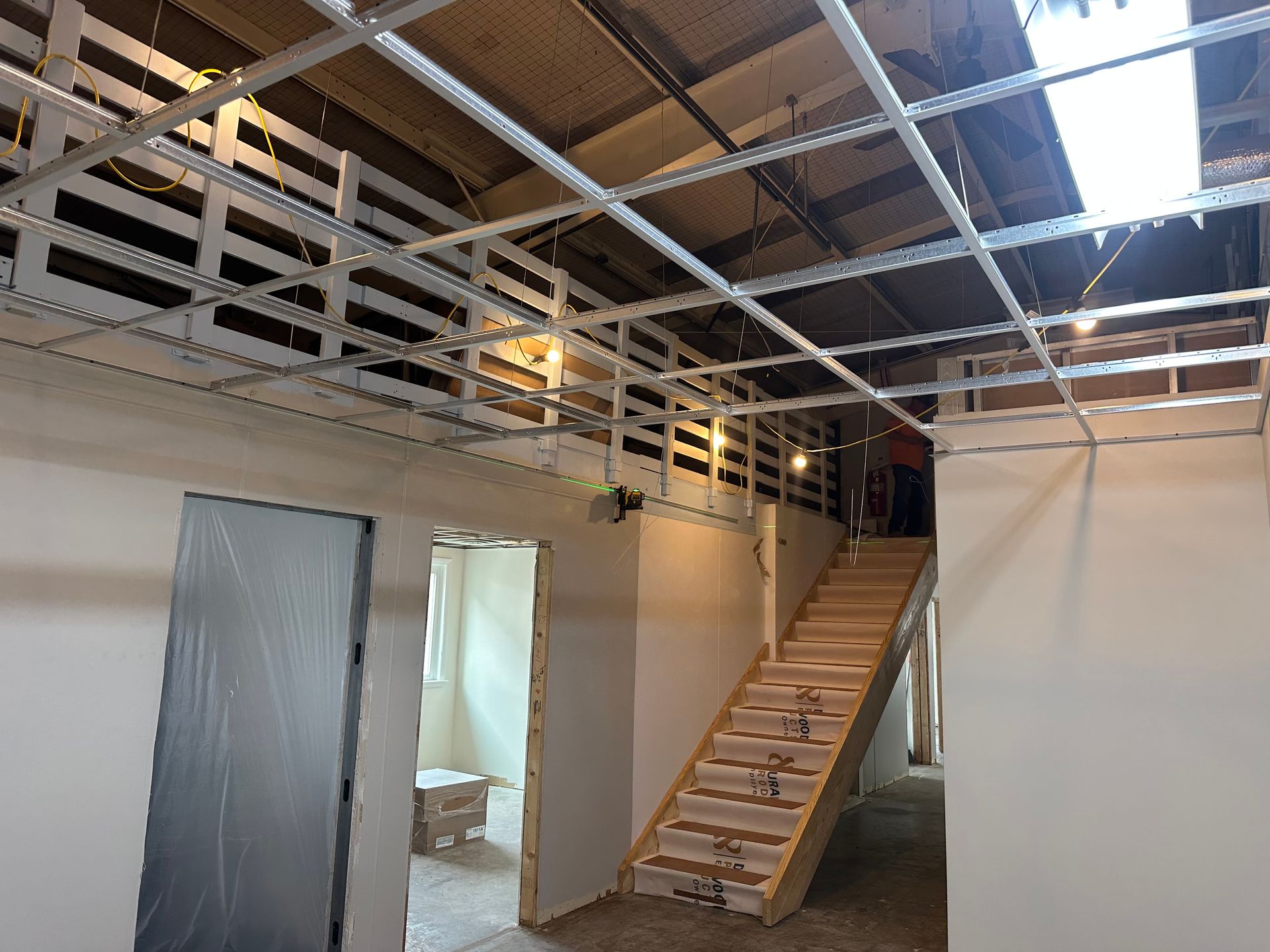 Interior view of a building under construction, featuring a staircase, exposed ceiling grid, and doorways.