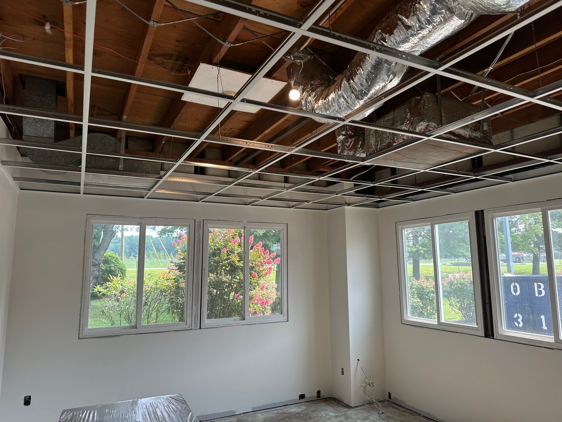 Interior view of a room under construction with exposed ceiling grid, windows, and painted walls.