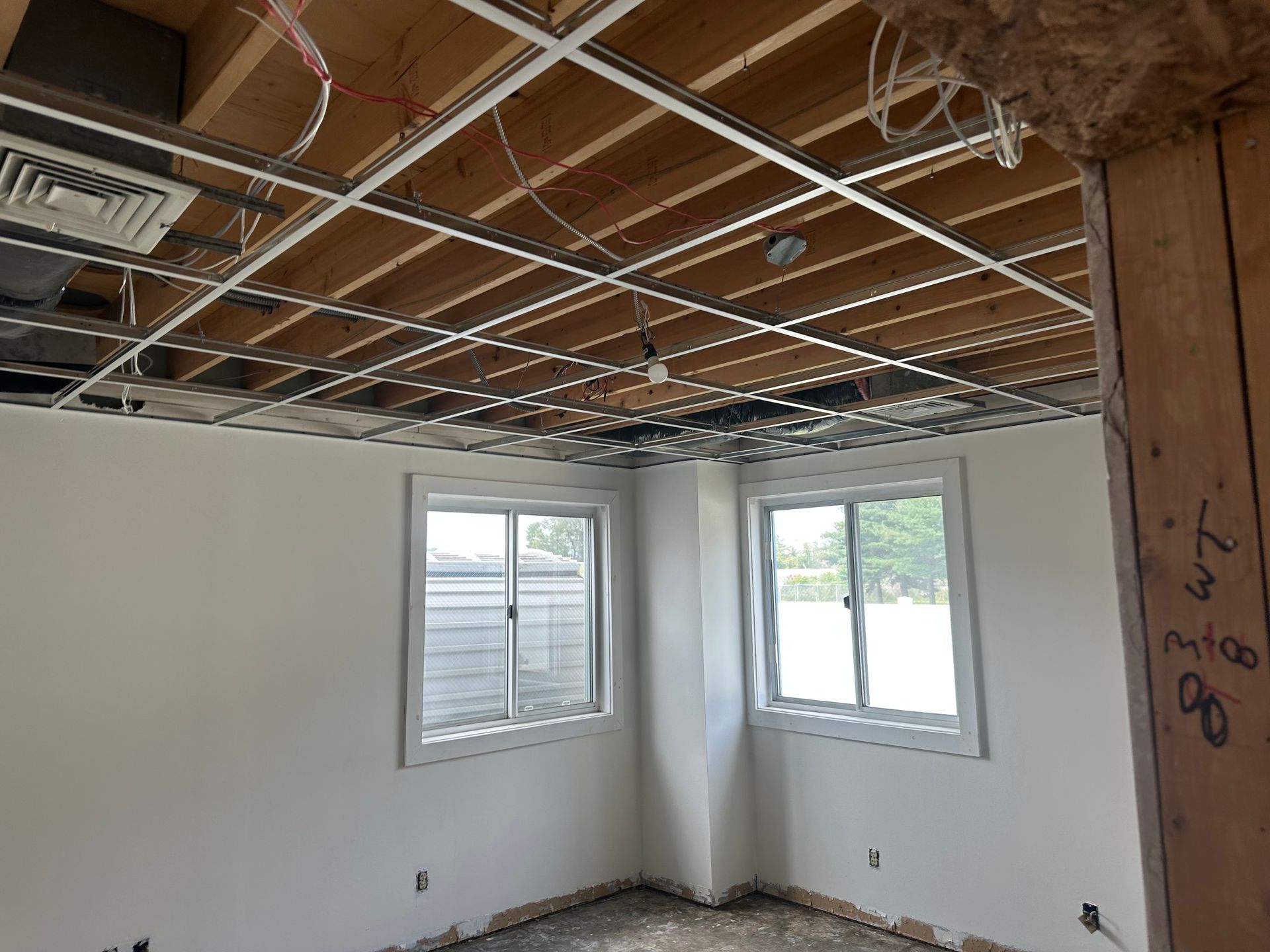 Interior room renovation with exposed ceiling grid, two windows, and unfinished walls.