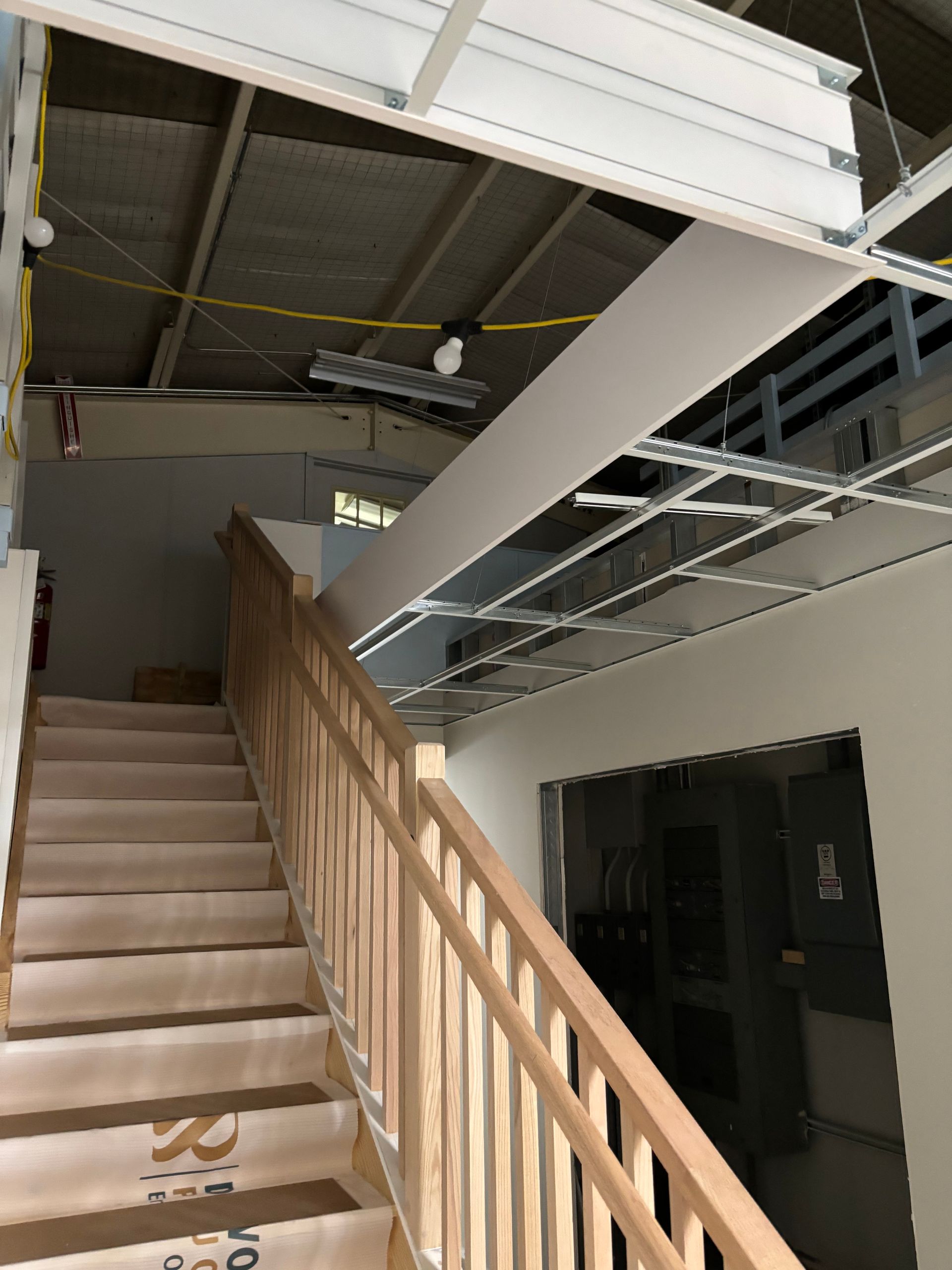 Wooden staircase inside a building. Handrail on the right, partial drywall ceiling.
