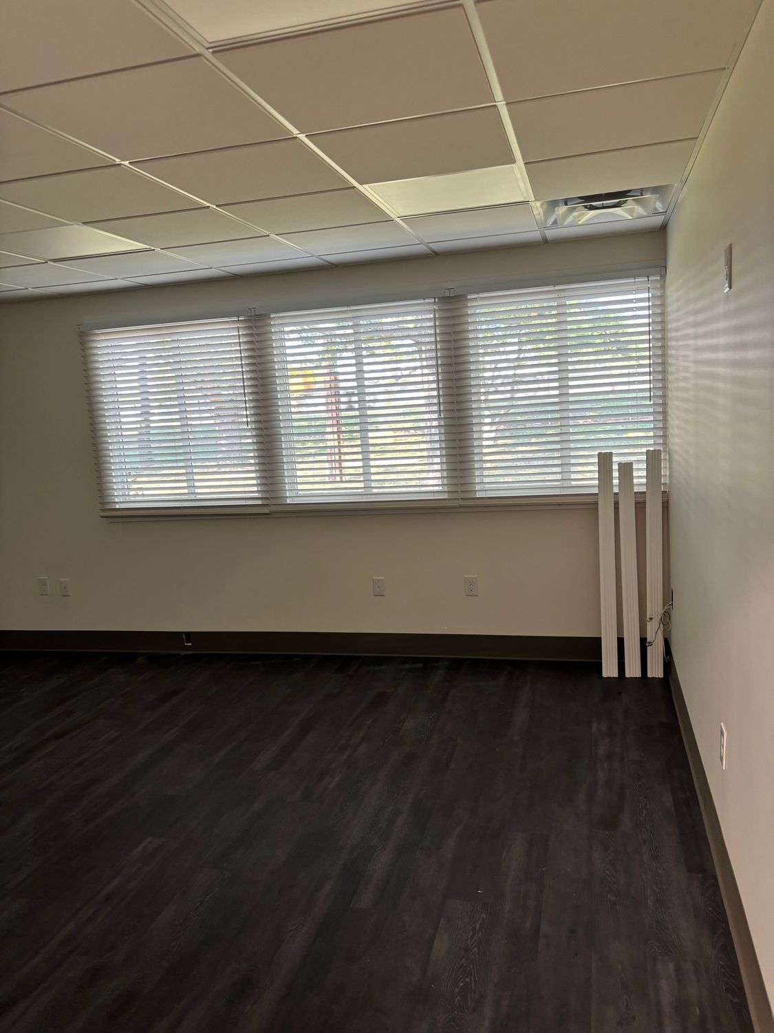 Empty room with three windows, white blinds, and dark wood-look flooring.
