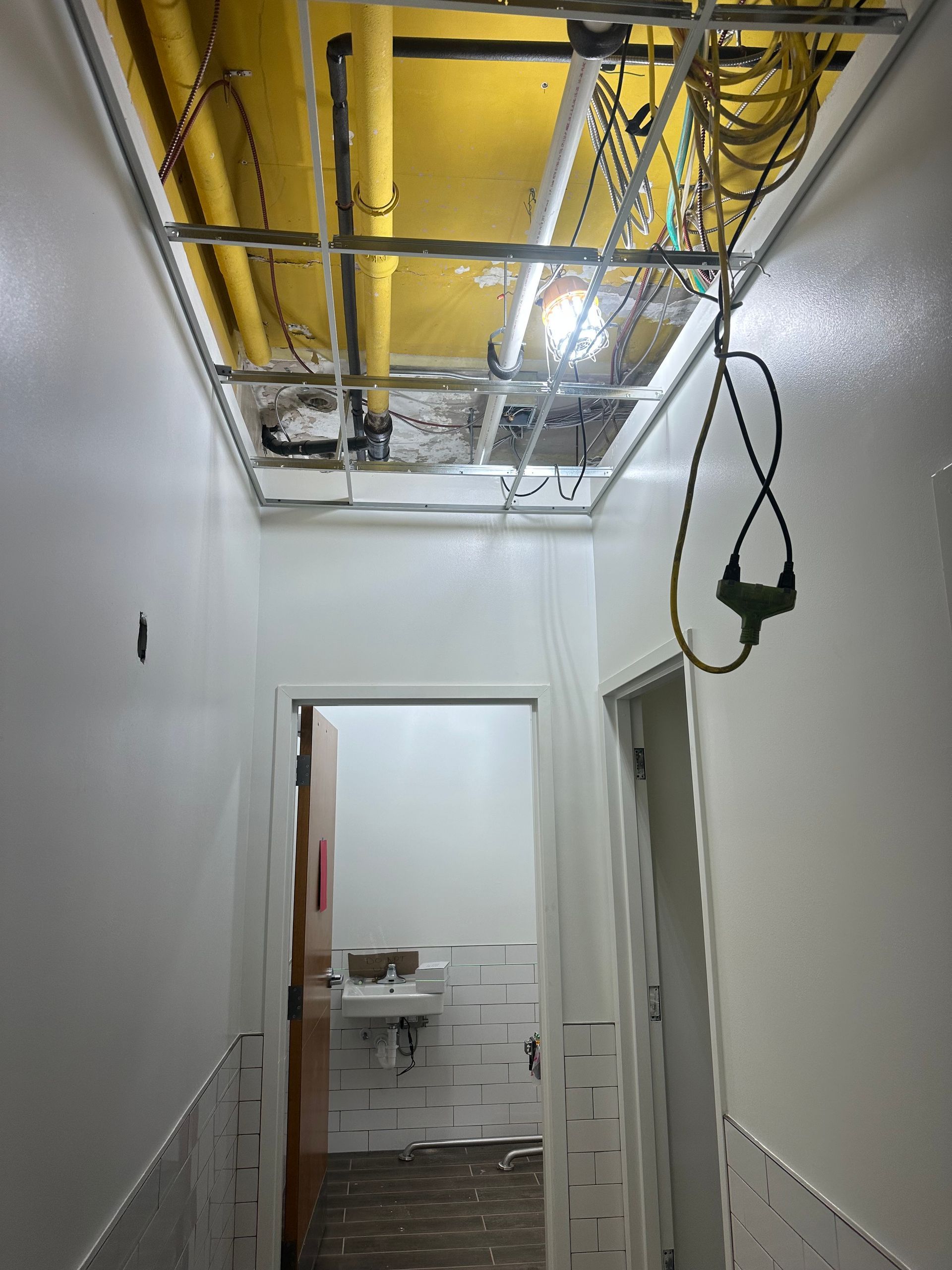 Hallway with exposed ceiling utilities, leading to a bathroom doorway. Walls are white; ceiling is yellow.