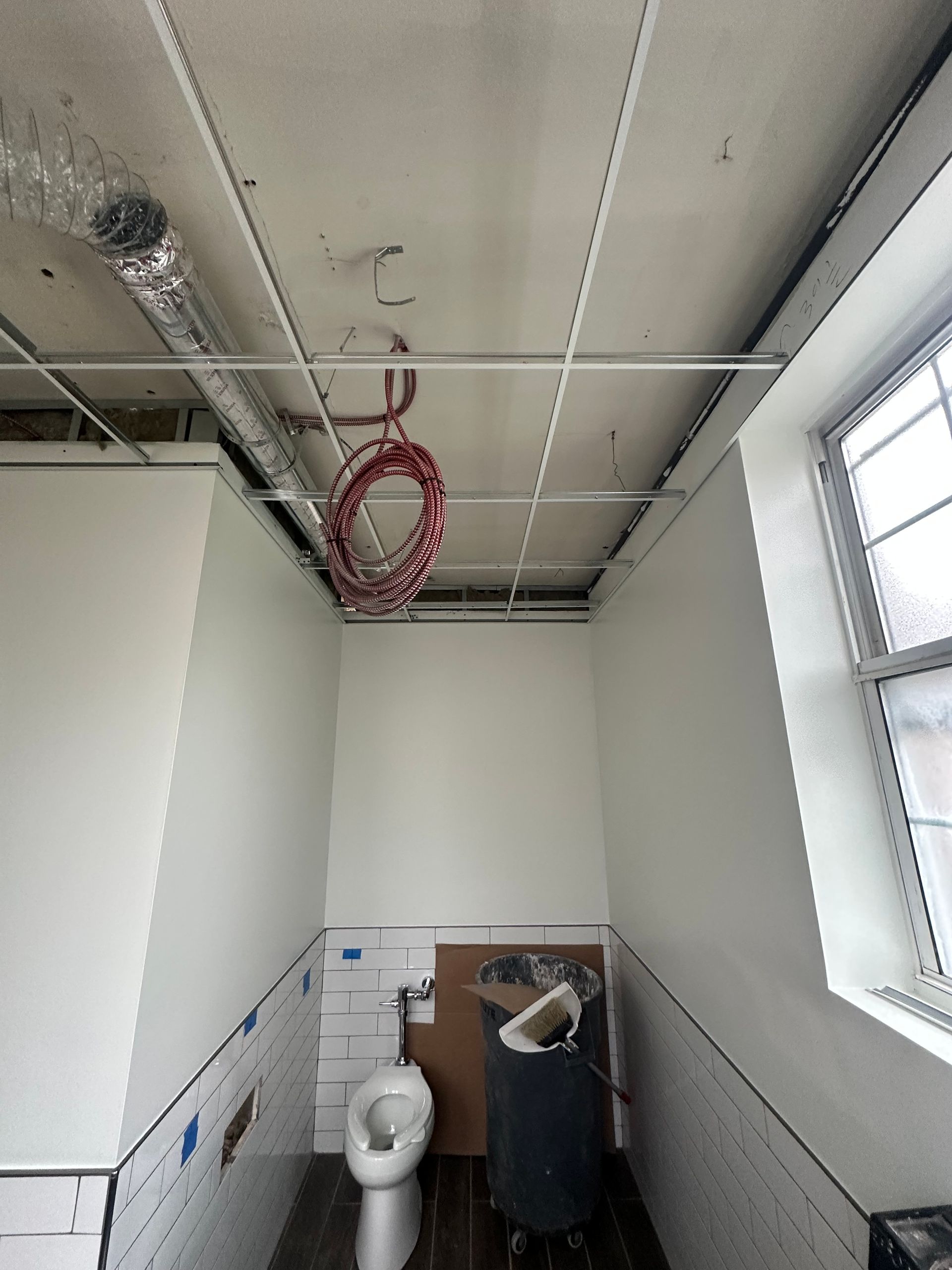 Small bathroom with exposed ceiling, toilet, and window on the right. Walls are white and partially tiled.