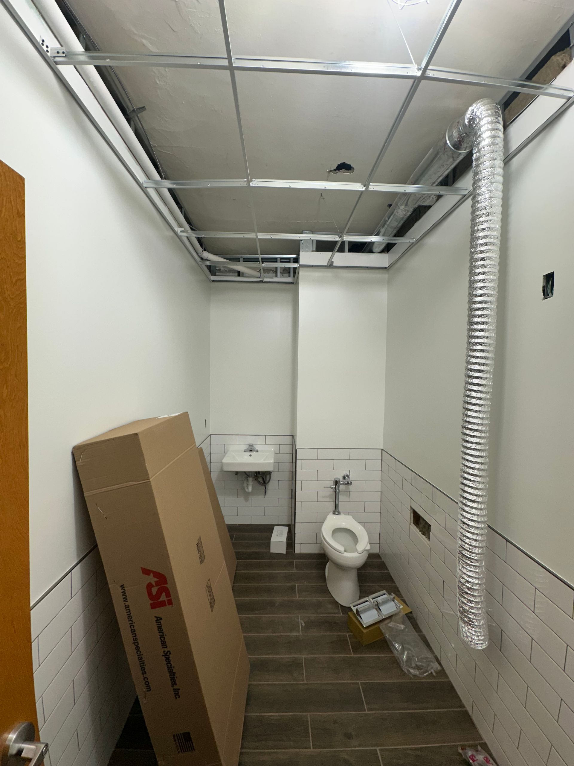 Bathroom under construction with toilet, sink, and ceiling grid, with ventilation duct. Box in foreground.