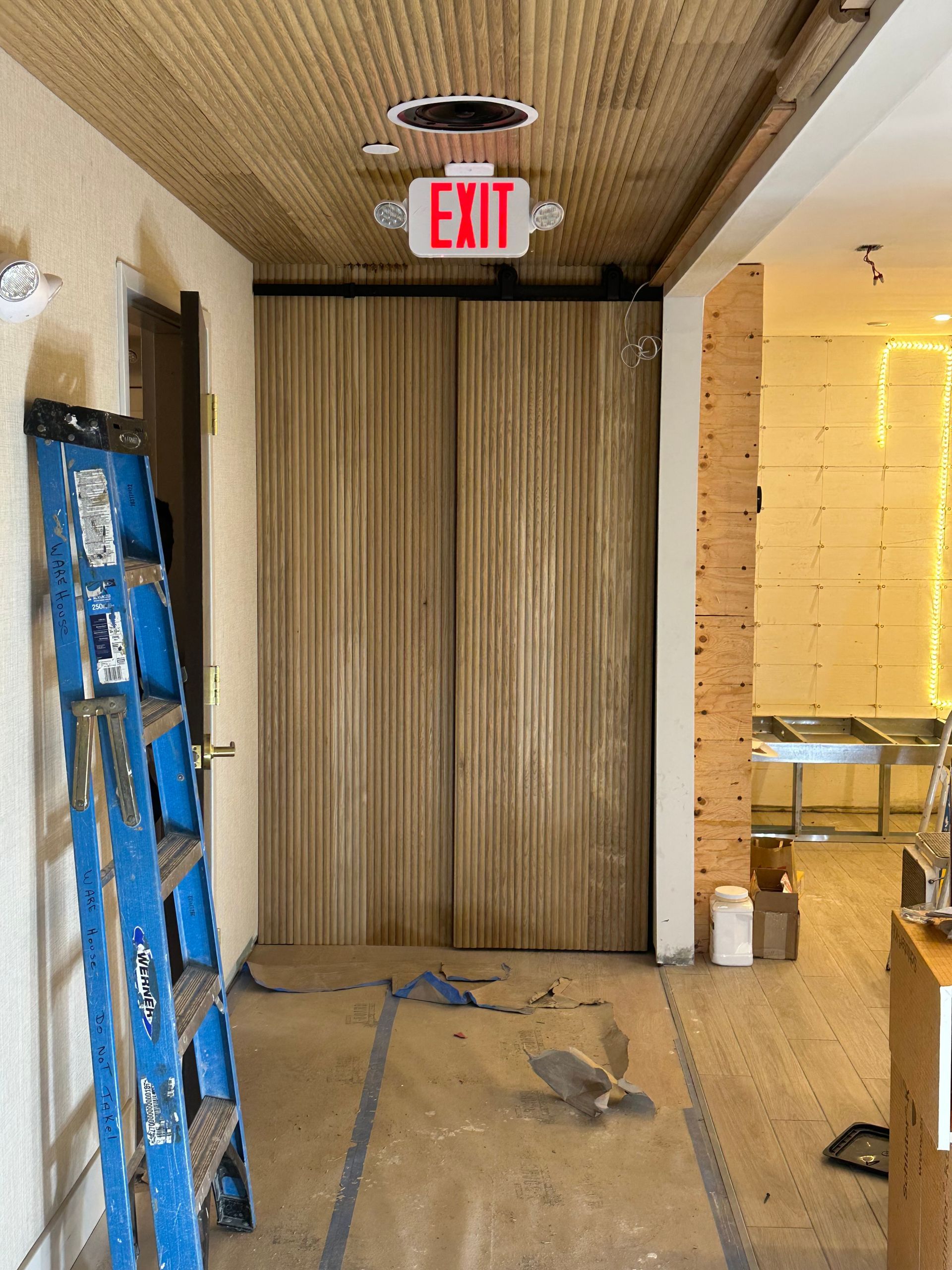 Hallway with exit sign, two closed doors, and a blue ladder.