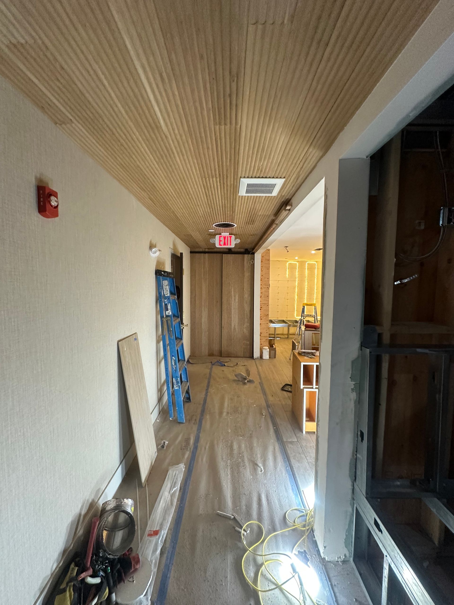 Hallway under construction, wood-slat ceiling, exposed framing, drywall, debris, blue tape on floor, exit sign.