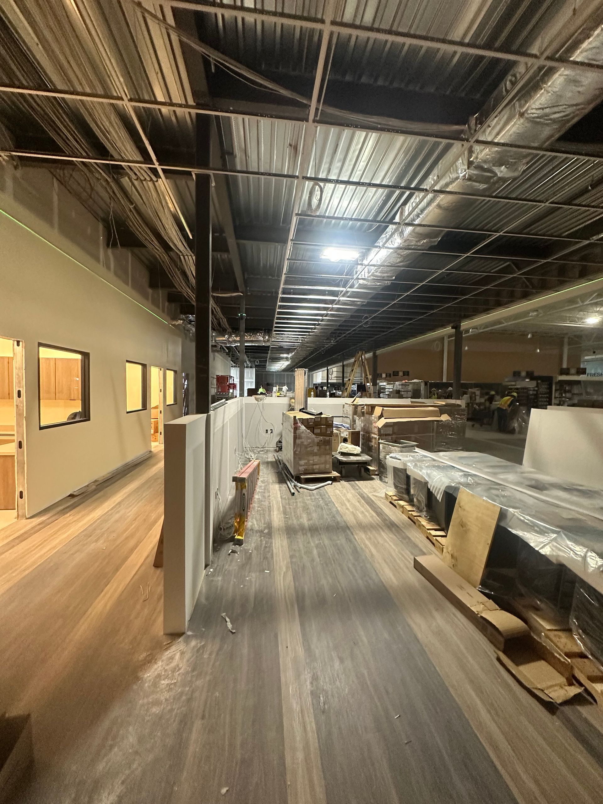 Interior view of a construction site. Long, warehouse-like space with exposed beams, drywall, and building materials.