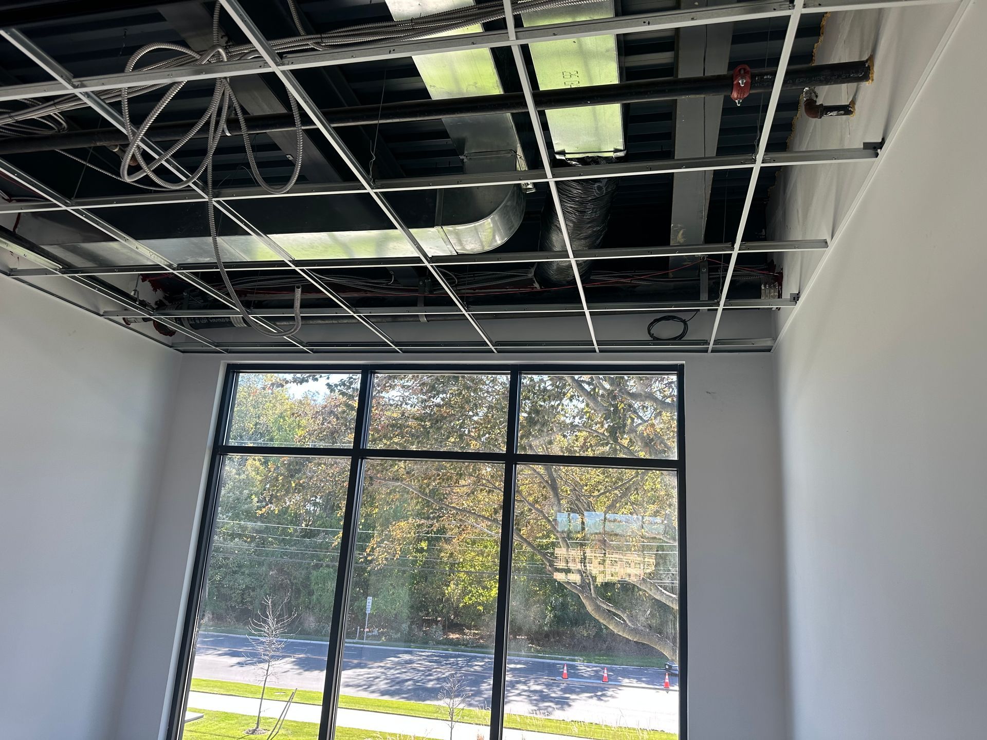 Interior room with exposed ceiling grid, HVAC, wiring, and large window overlooking trees.