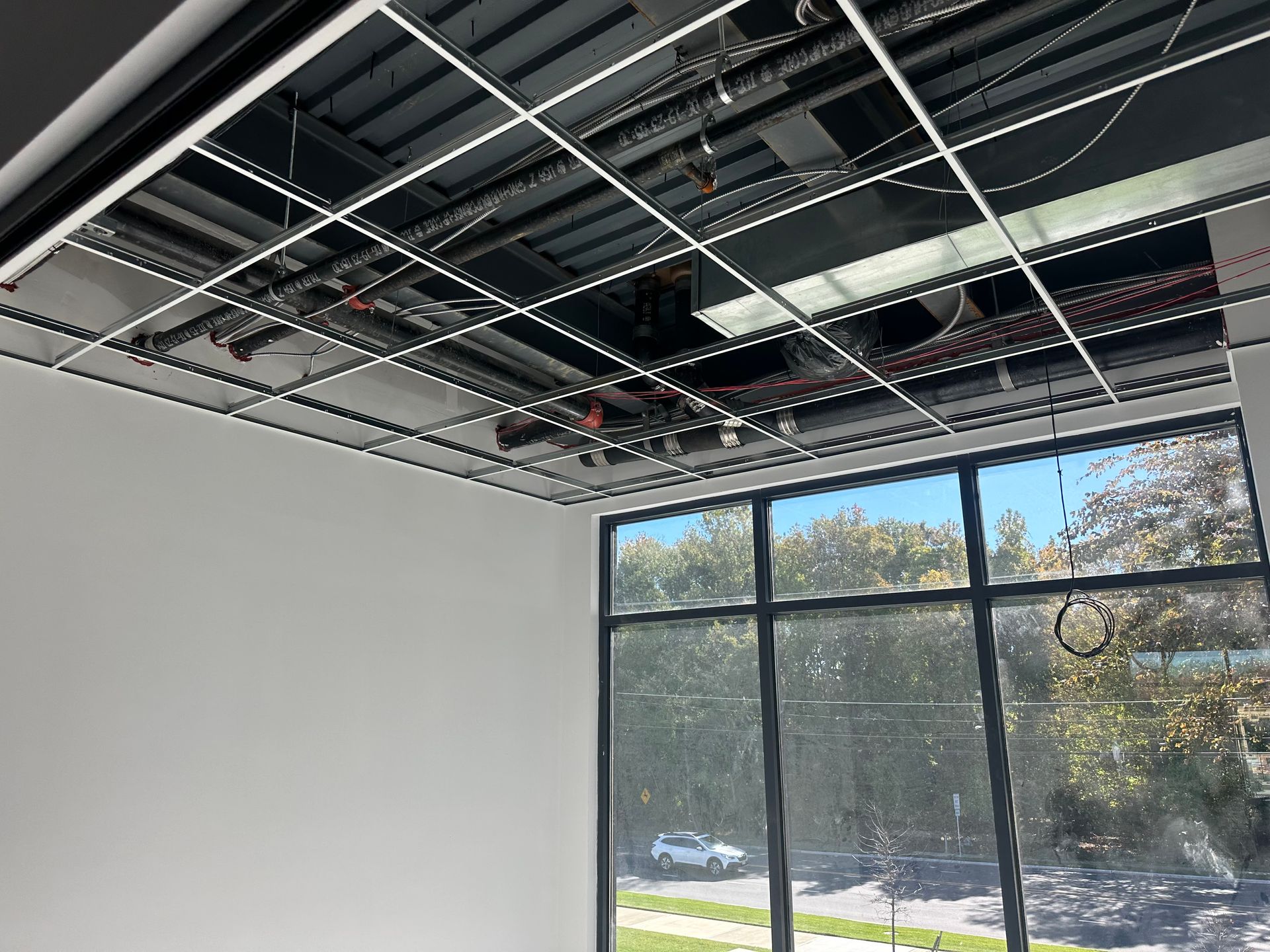 Open ceiling with metal grid and visible pipes, wires, and ductwork above a room with large windows and a view of trees.