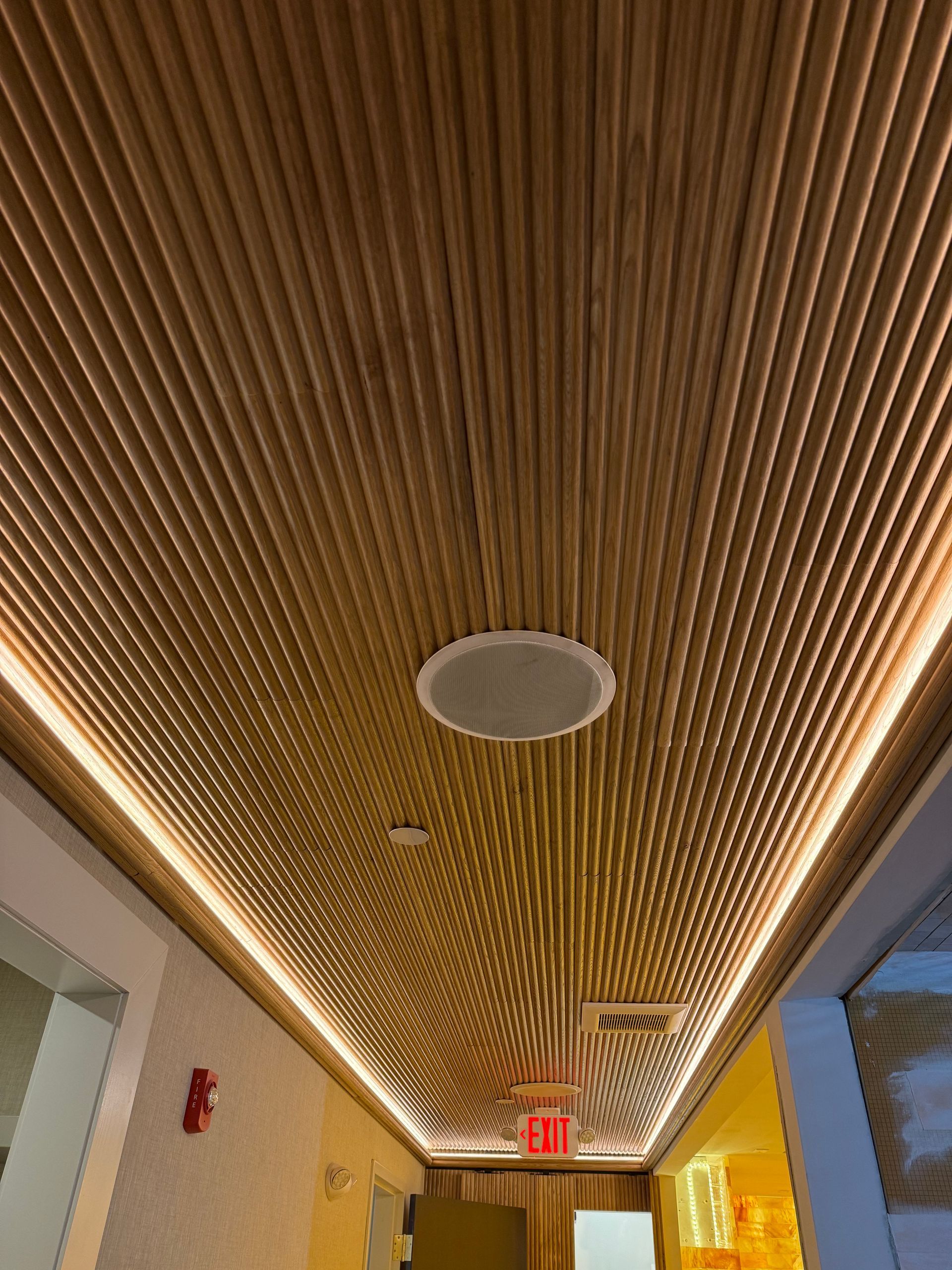 Wooden slatted ceiling with recessed lighting and a speaker. Hallway with exit sign and red fire alarm.