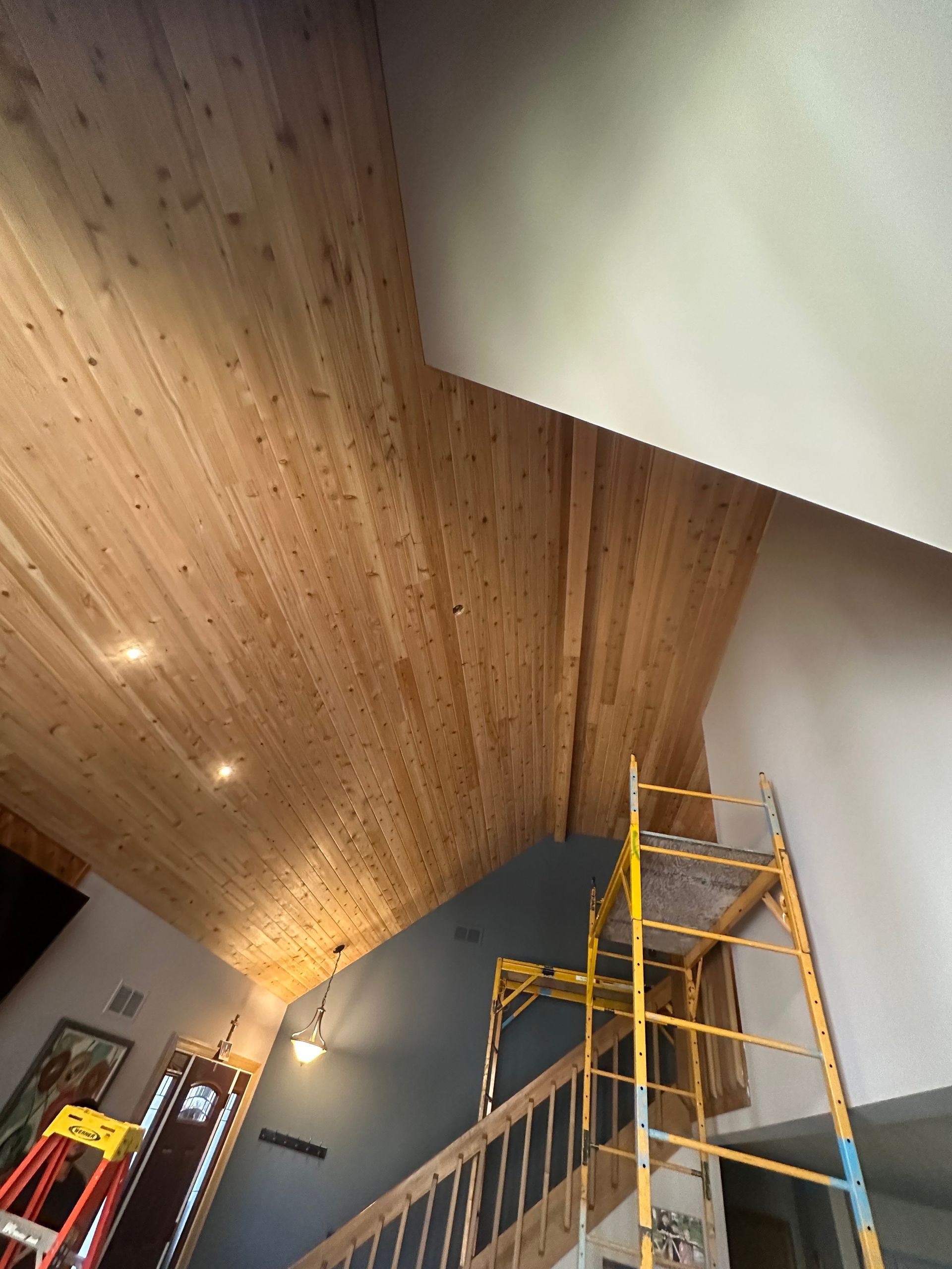 Wooden plank ceiling with recessed lights, against a light-colored wall with a yellow scaffolding.