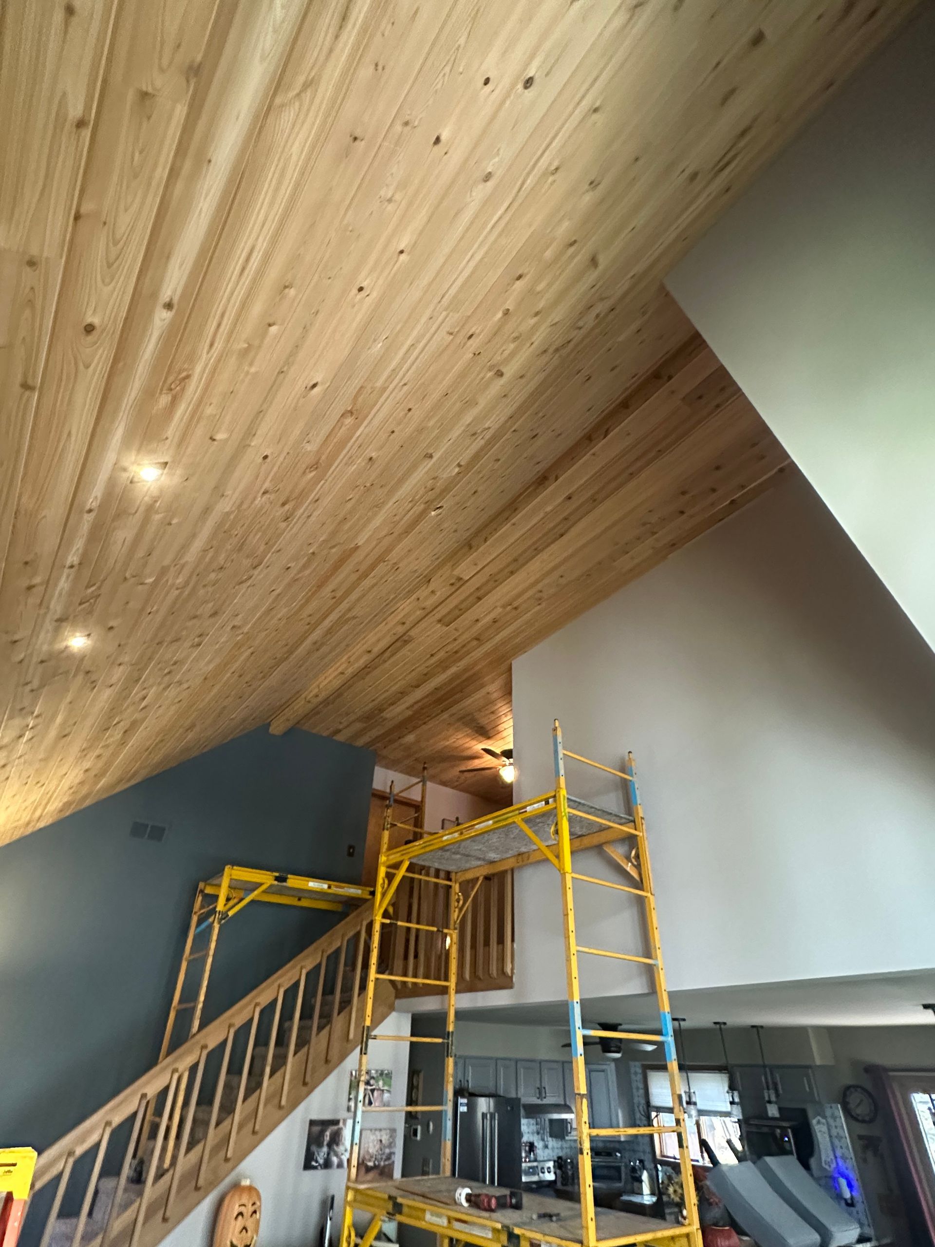 Wooden ceiling installed in a room with a tall, angled wall. Yellow scaffolding supports the installation.