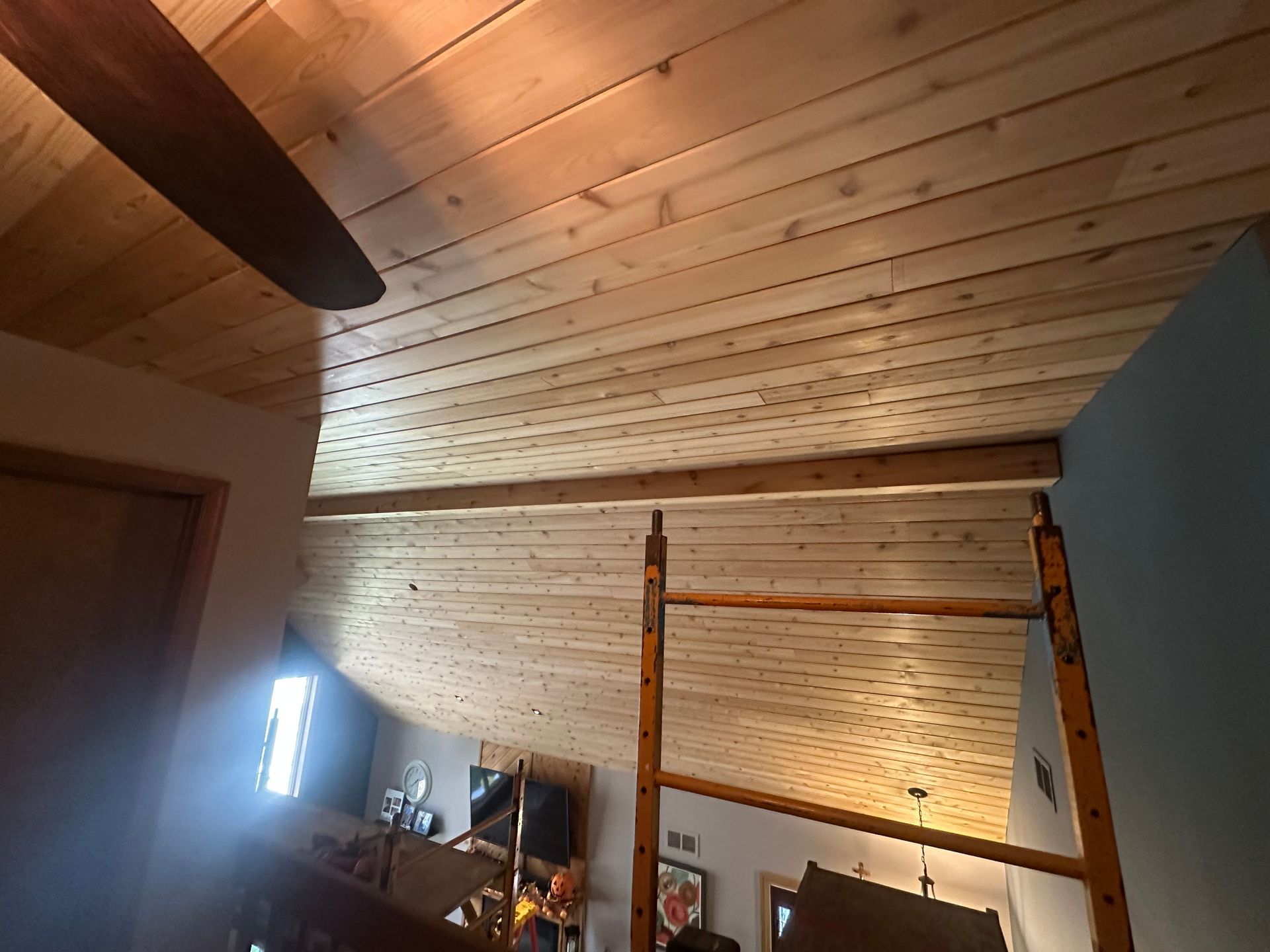 Wood-paneled ceiling with a fan blade and a wooden scaffolding setup in a room, with a glimpse of a living area.