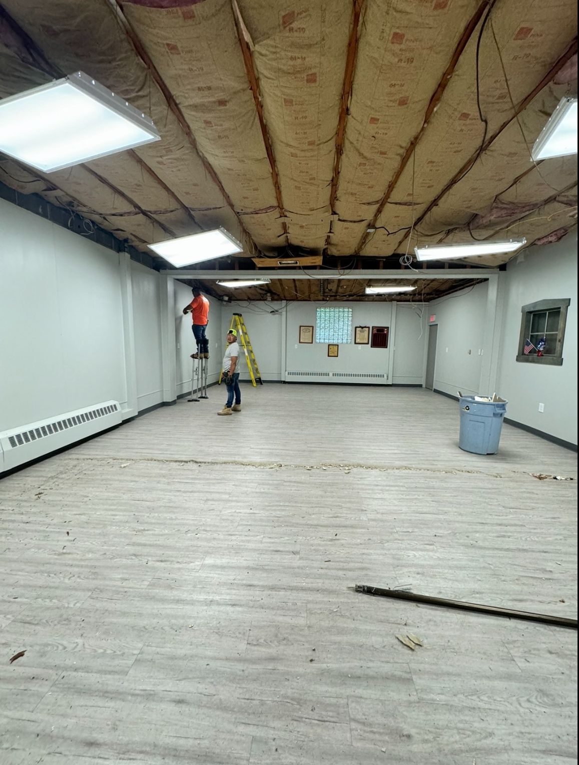 Room under renovation; ceiling insulation exposed, workers on ladders, white walls, gray flooring.