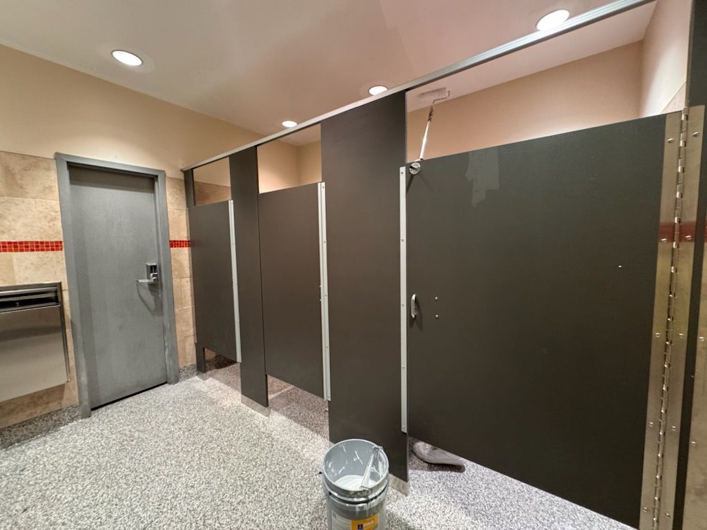 Public restroom with three stalls and a gray door. Flooring is speckled. A bucket sits in one stall.