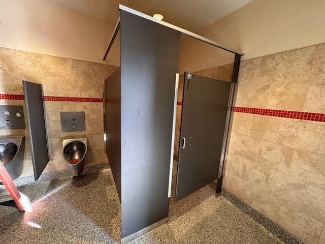 Public restroom with dark gray stalls, tan tile walls, and a urinal.
