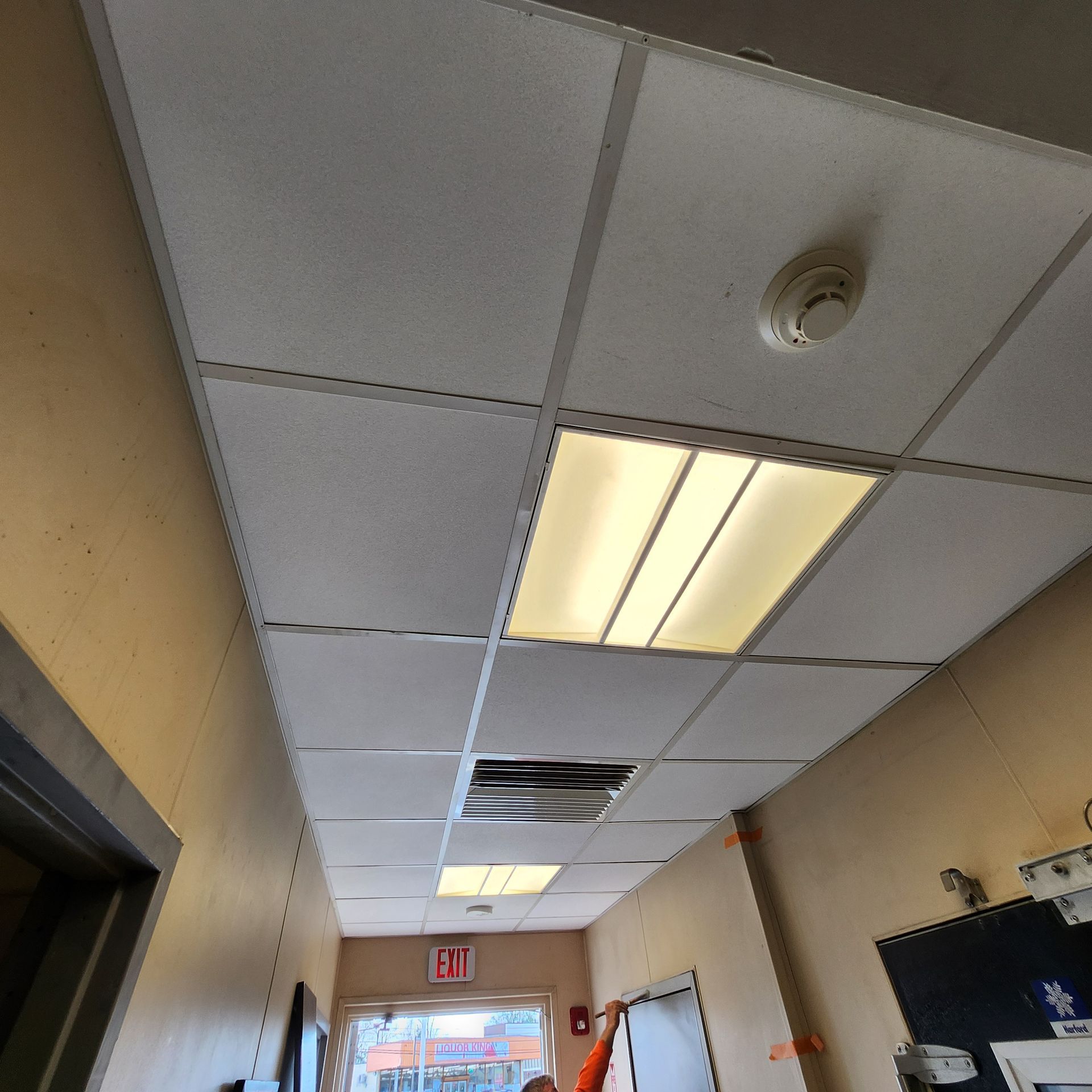 Low-angle view of a hallway ceiling with ceiling tiles, lights, and a smoke detector.