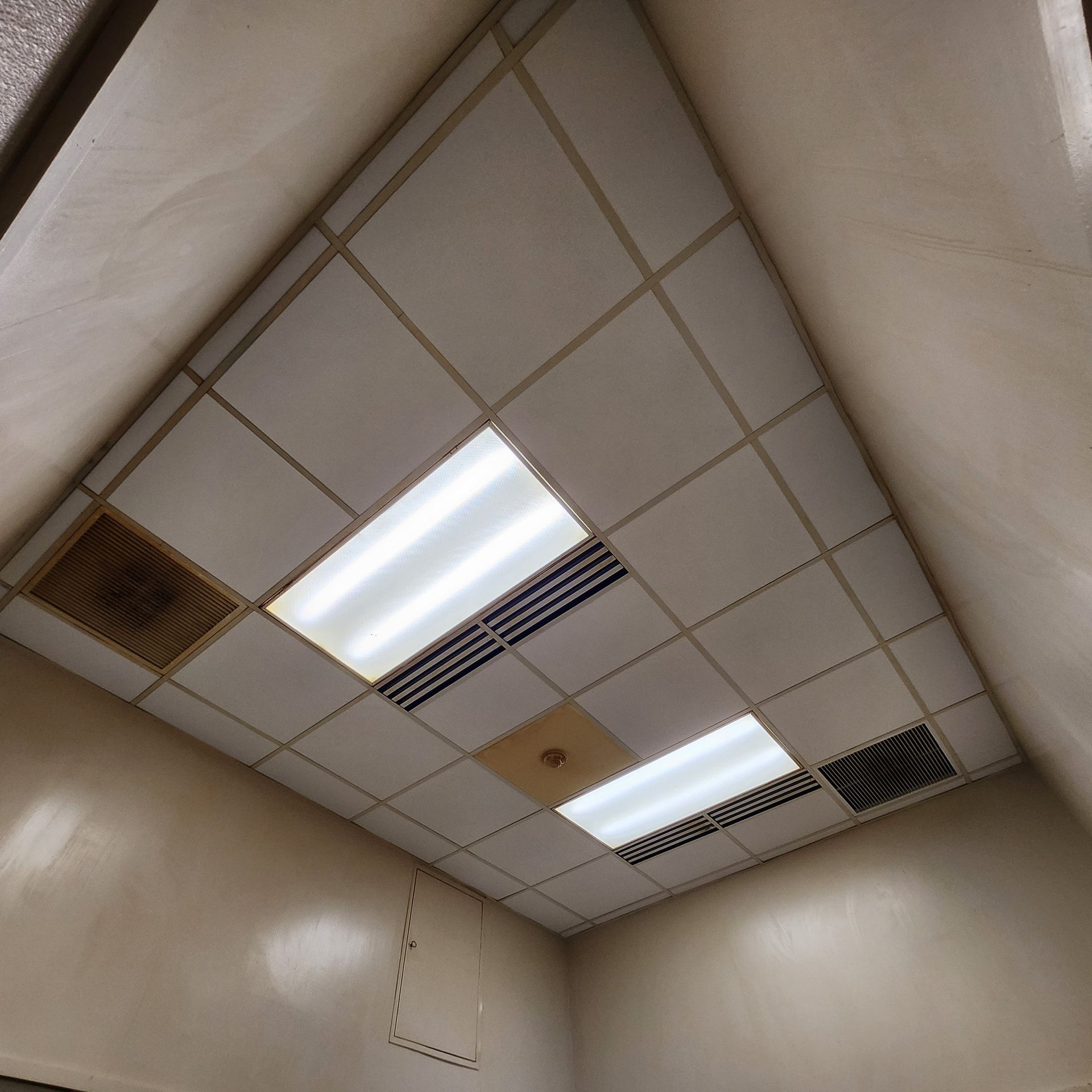 Overhead view of a tiled ceiling with fluorescent lights and vents. Beige walls surround.