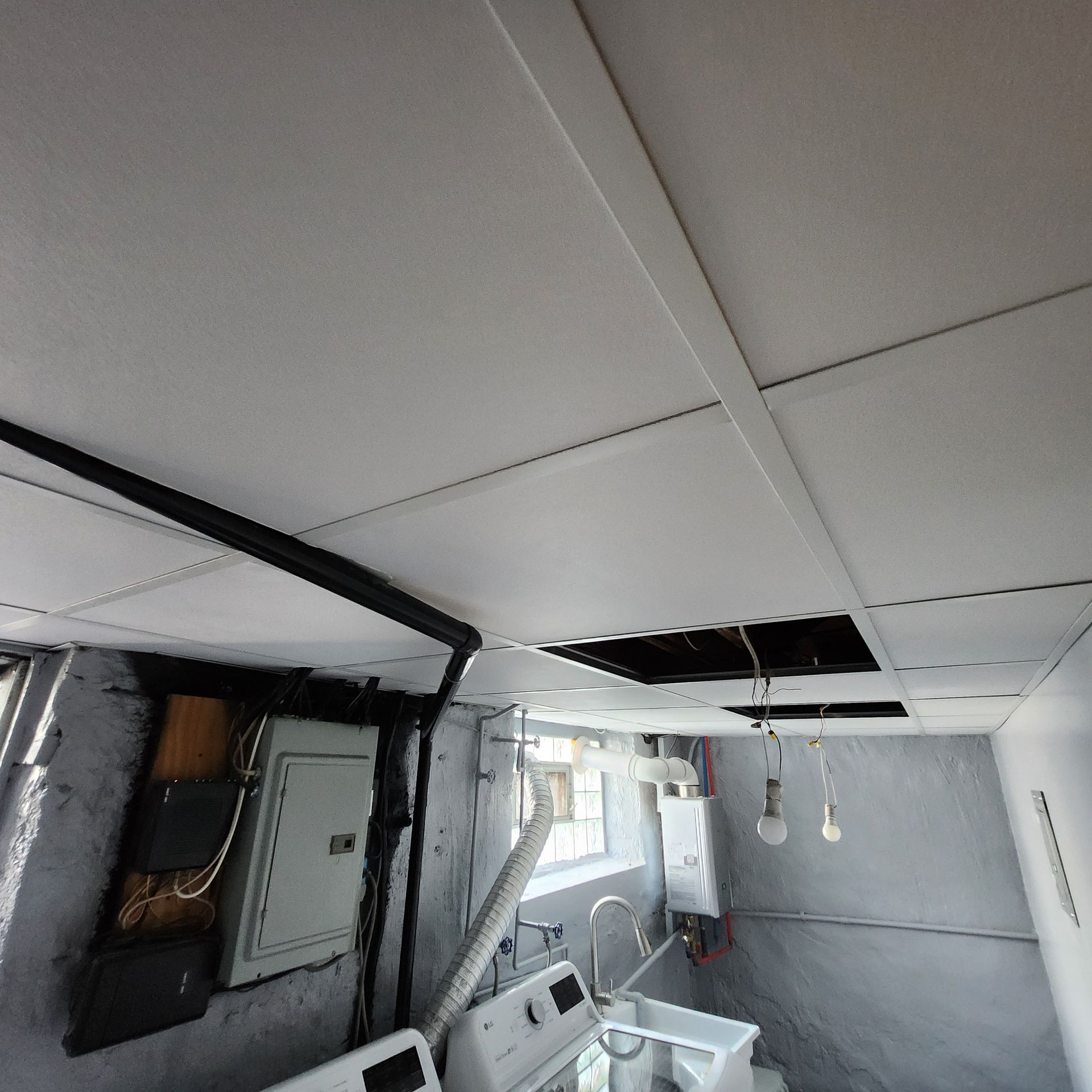 Ceiling tiles in a basement with electrical panel, exposed wires, and a washing machine.