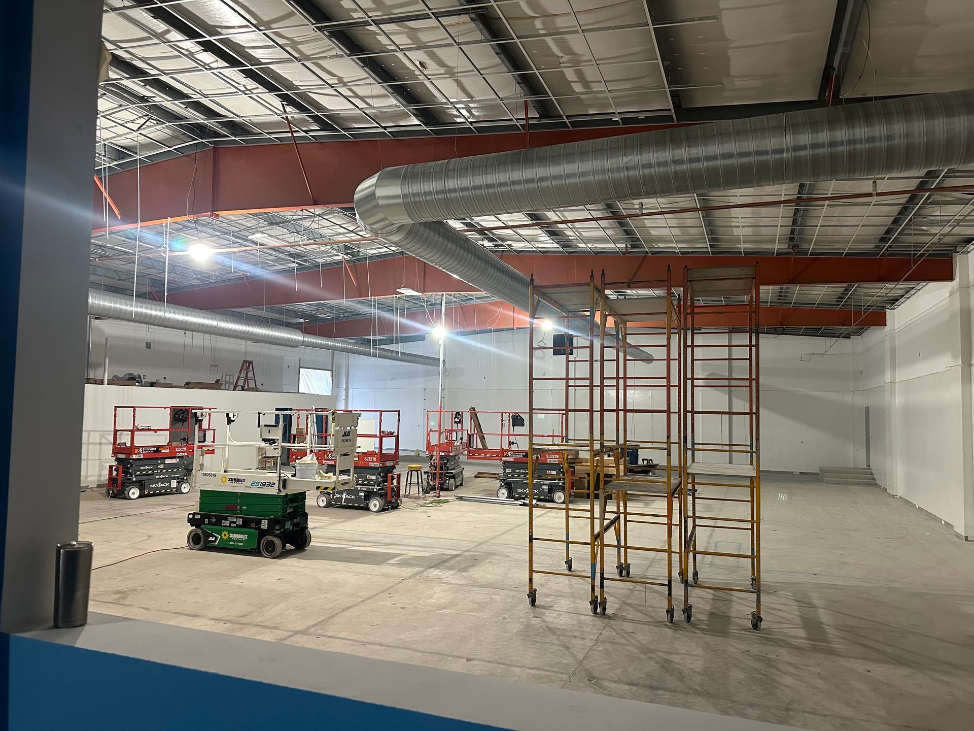 Construction site interior with equipment like lifts and scaffolding. Exposed ceiling, white walls, and concrete floor.
