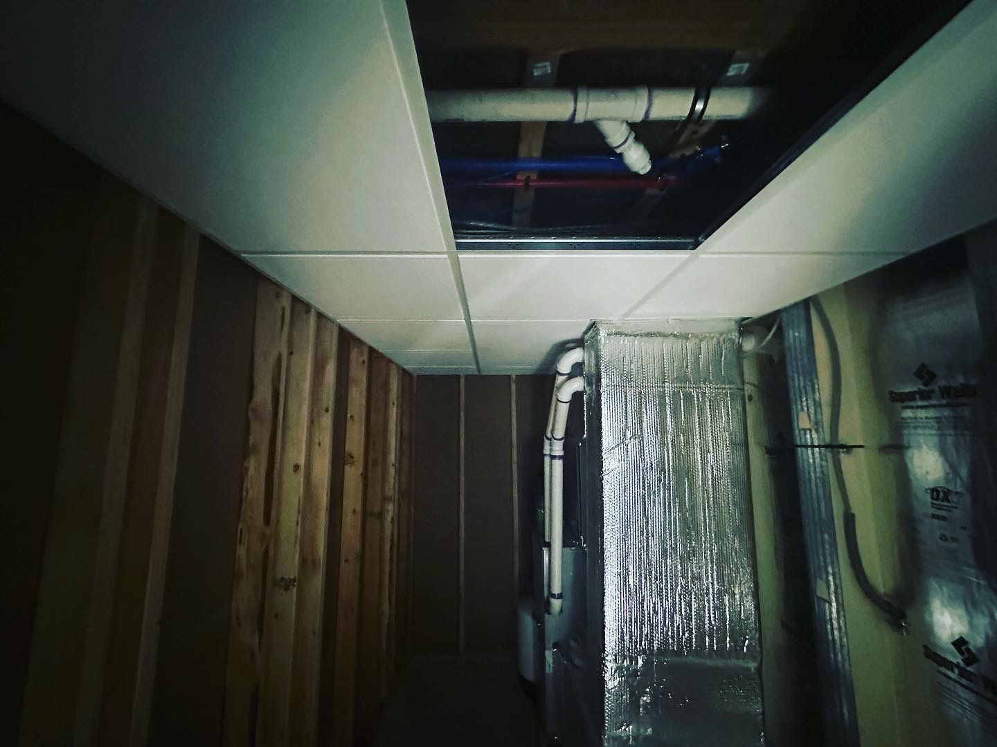 Basement ceiling with open section revealing pipes and ductwork, surrounded by wood paneling and insulation.