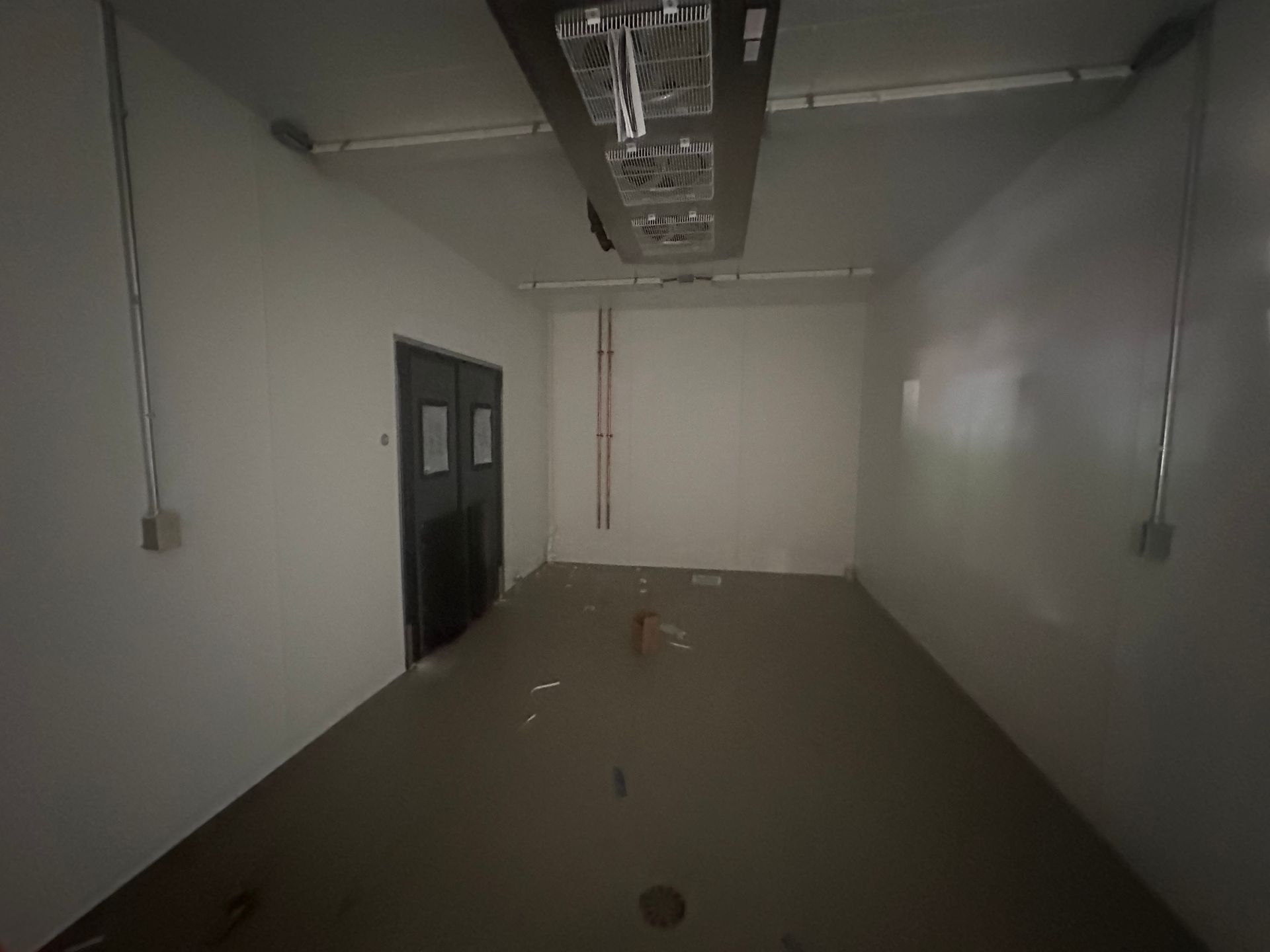 Empty, white-walled room with a dark door, conduit, and ceiling-mounted equipment. Floor is light brown.