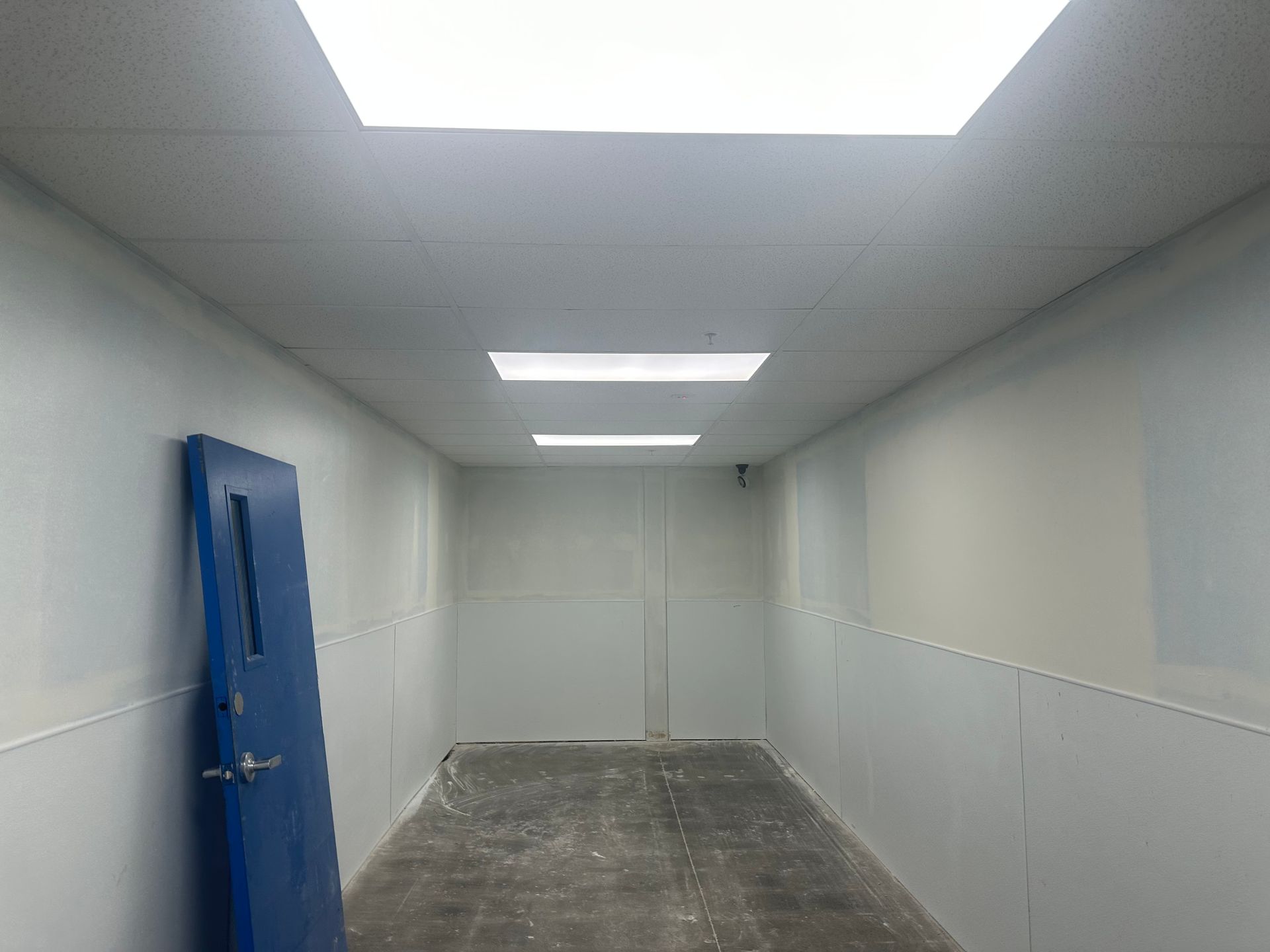 Long, white hallway with blue door on left and rectangular ceiling lights.