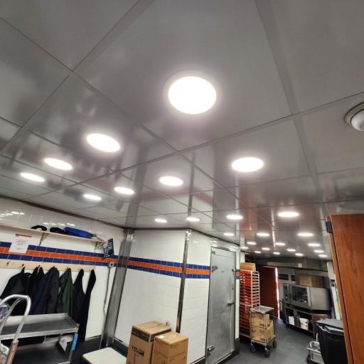 A commercial kitchen interior with bright overhead lights, stainless steel surfaces, and work areas.