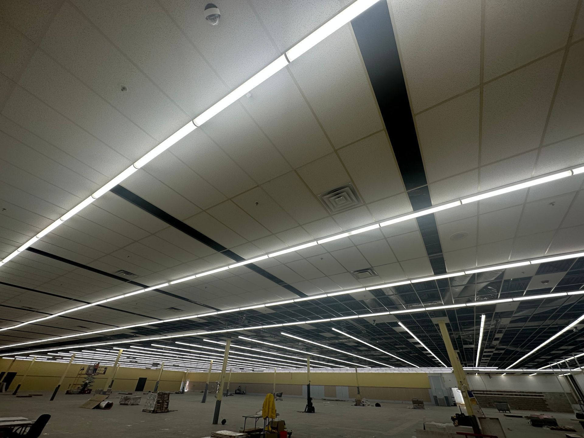 Low-angle shot of an unfinished interior space, with a grid ceiling, long fluorescent lights, and construction equipment.