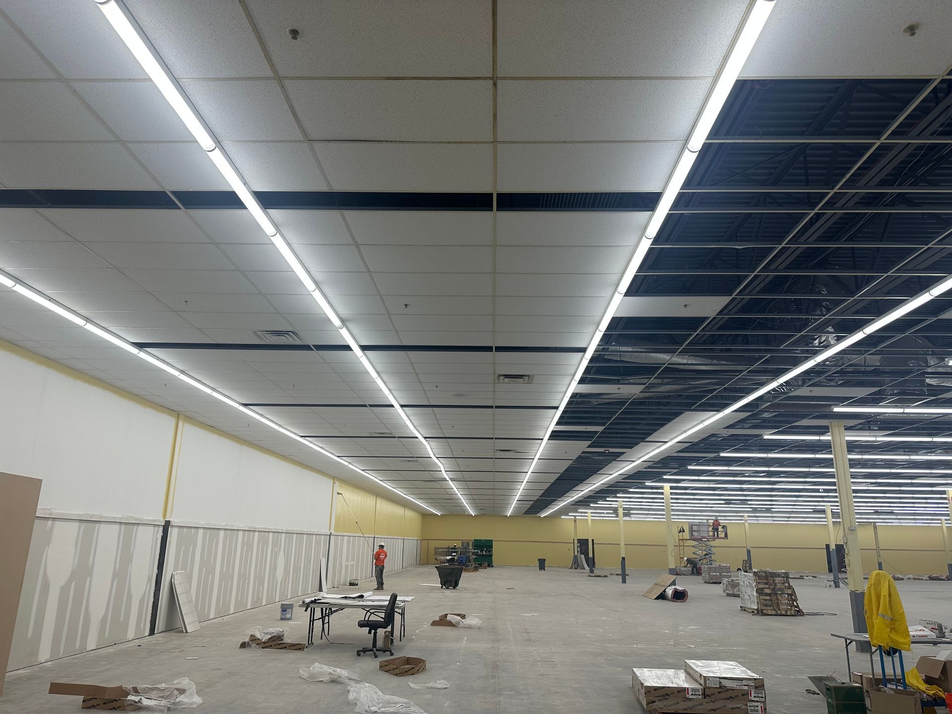 Construction site interior with fluorescent lights, exposed ceiling, and scattered materials.