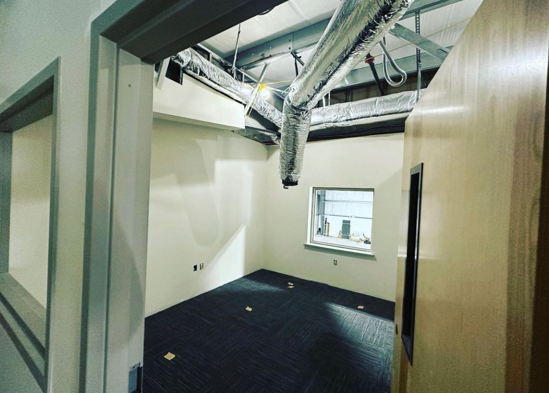 Empty room with open ceiling showing ductwork, window, and doorframes. Dark carpet, beige walls.