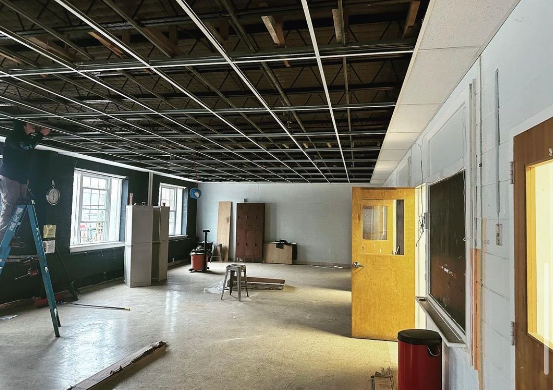 Interior room under construction, exposed ceiling grid, debris on floor, doors and windows visible.