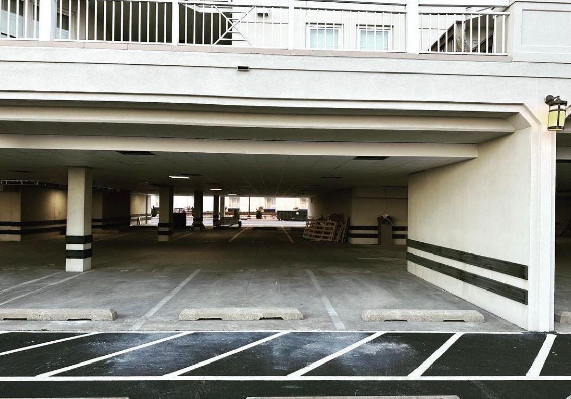 Empty, covered parking garage beneath a building; concrete and support beams; daylight.