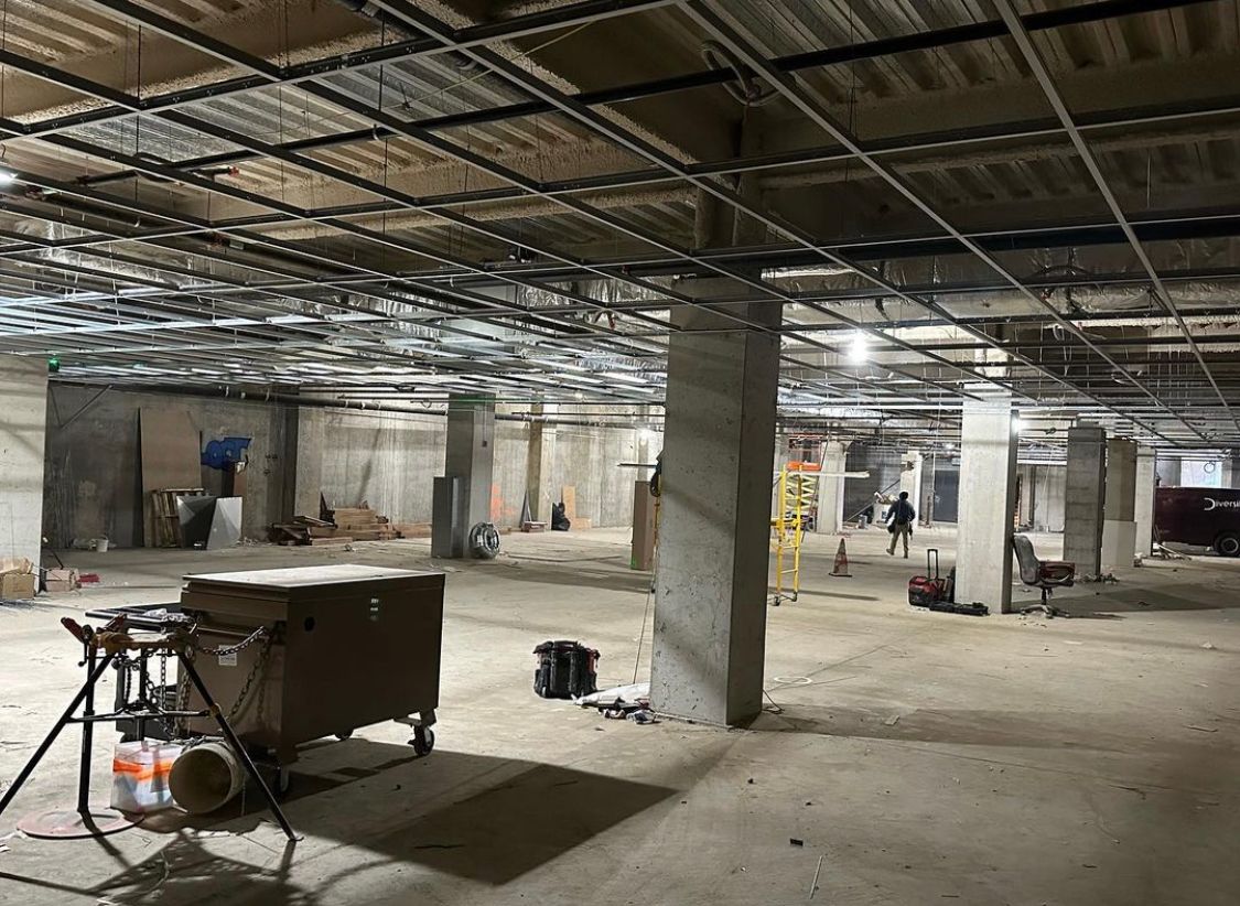 Construction site: unfinished interior with concrete pillars, exposed ceiling, and tools. Workers in the distance.