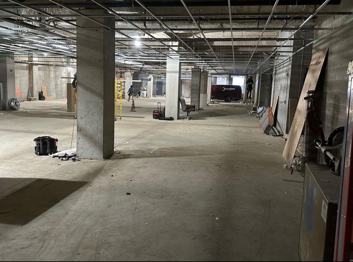 Interior of a concrete building in disrepair, exposed ceiling infrastructure, debris on the floor, some daylight.