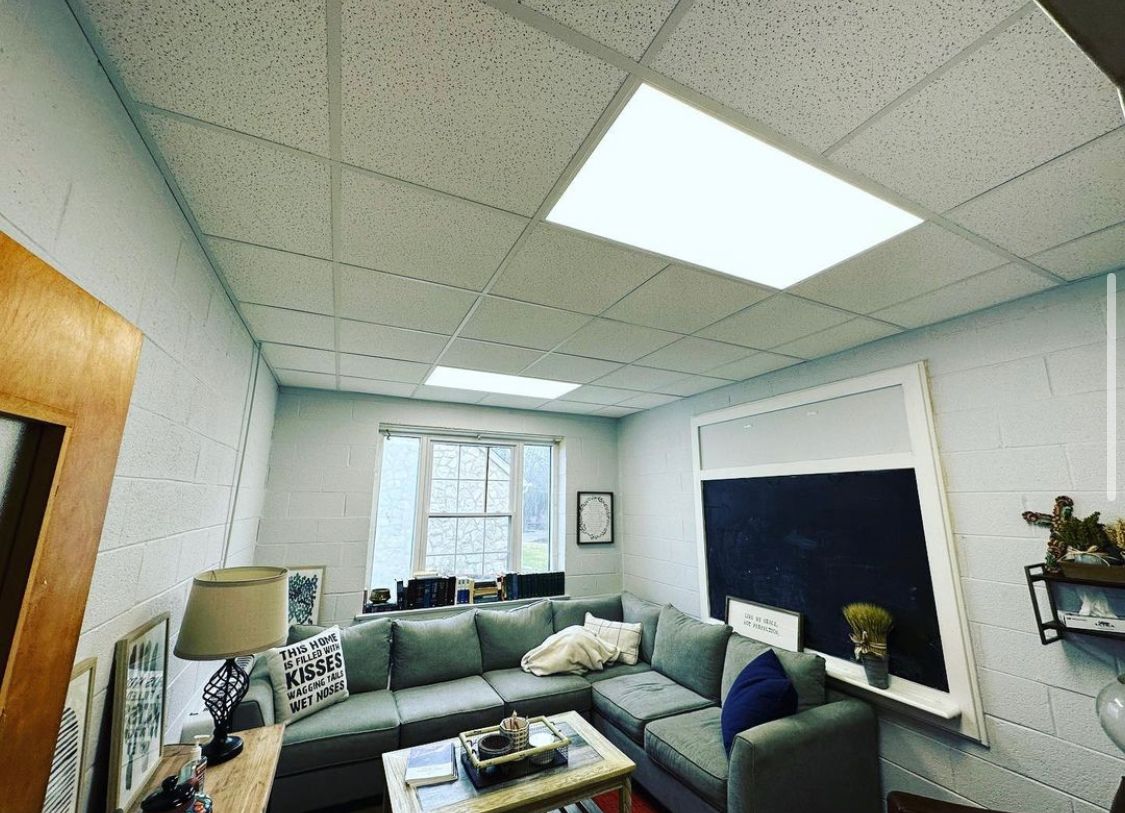 Cozy office with gray sectional, small window, black chalkboard, and ceiling lights.