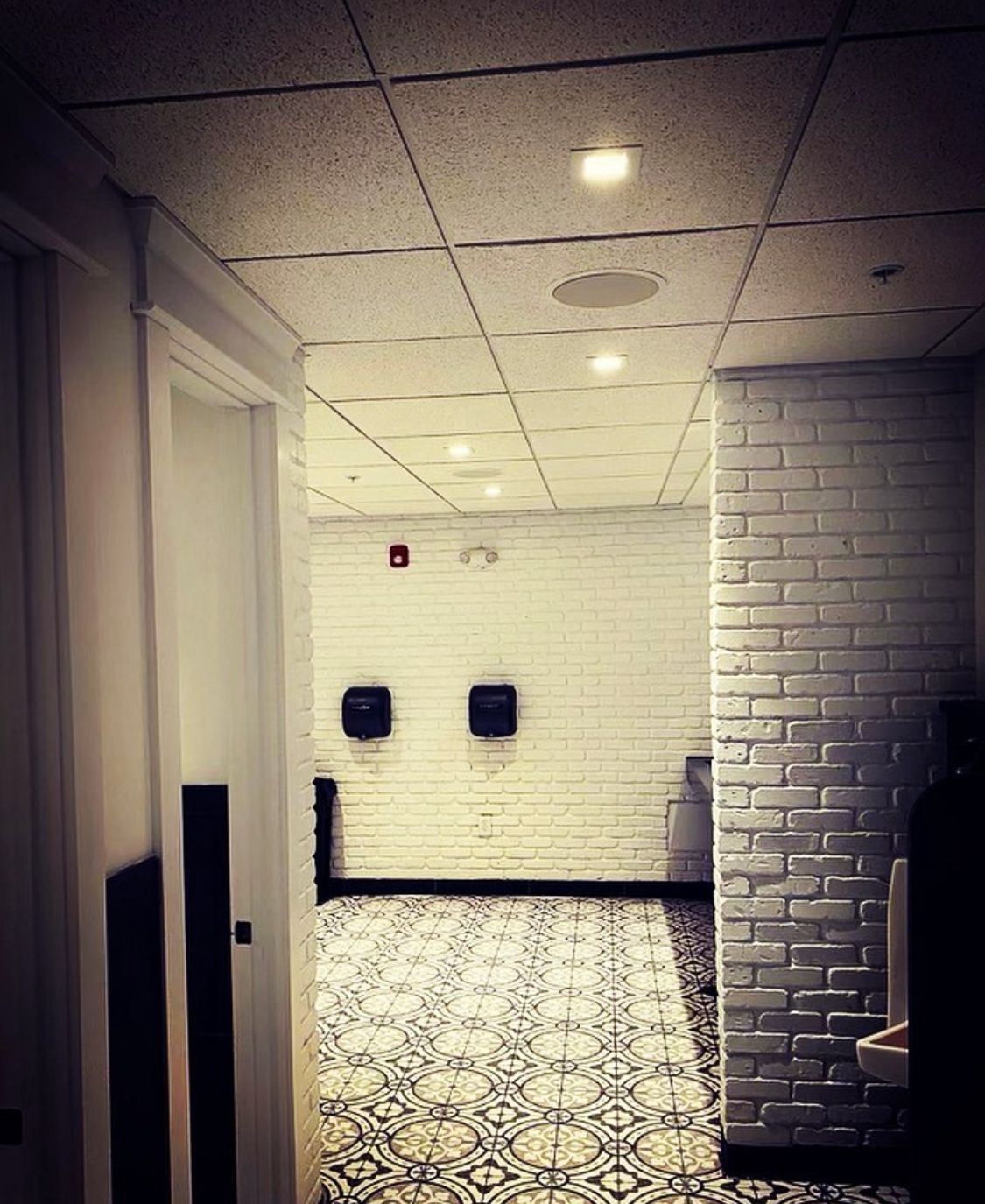 Hallway with white brick walls, patterned floor tiles, recessed ceiling lights, and two soap dispensers.