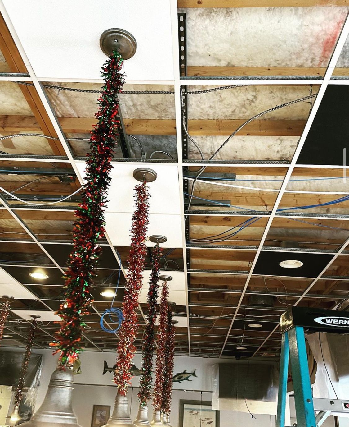 Lights with tinsel hang from a ceiling with missing panels, a ladder on the side.