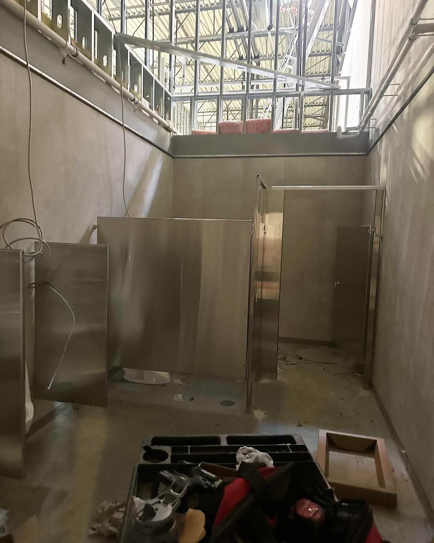 Construction site, restroom stalls in progress. Unfinished walls, metal stalls, tools on floor, exposed ceiling beams.