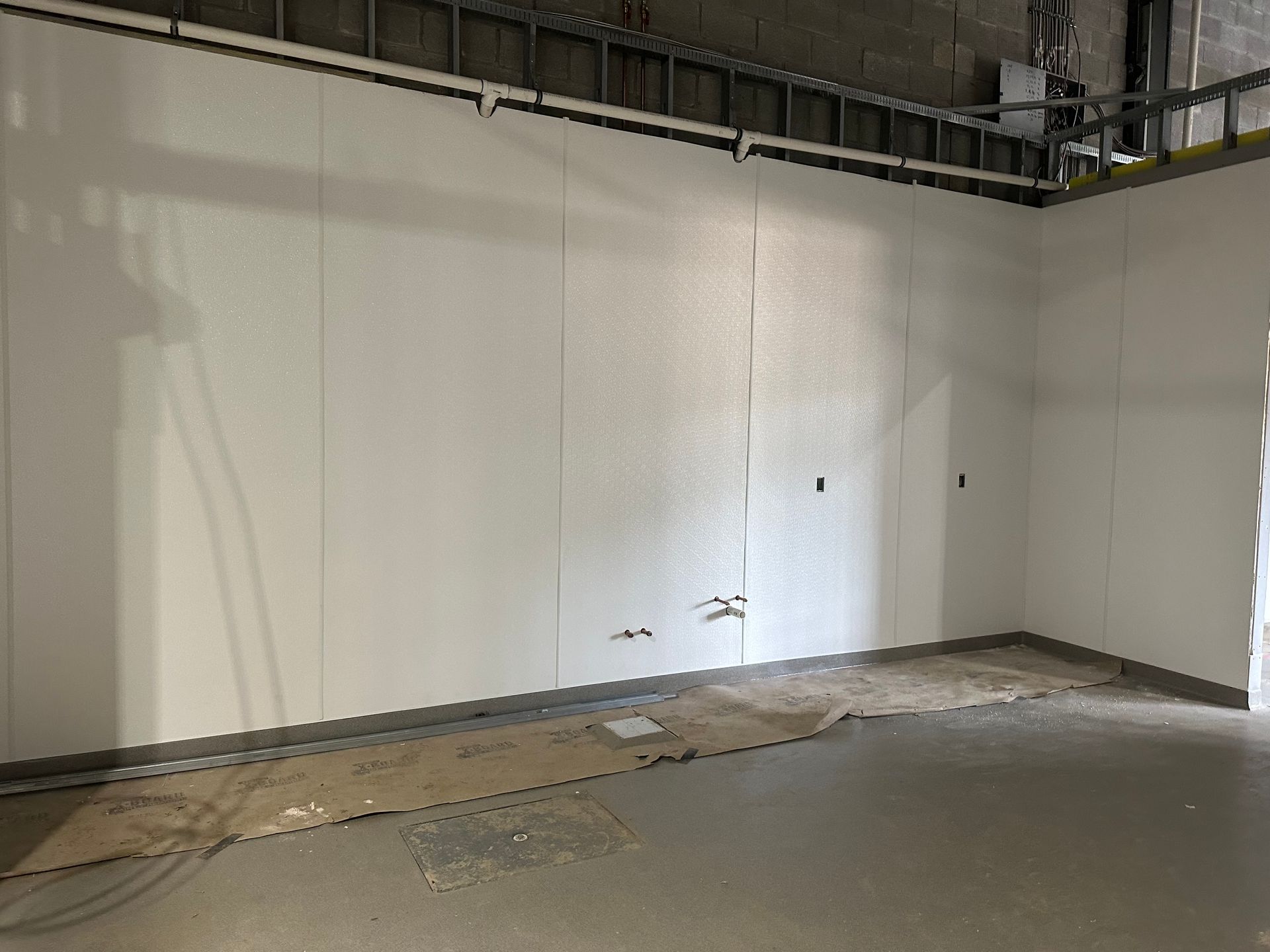 Interior wall with white panels, unfinished floor, and exposed ceiling structure.