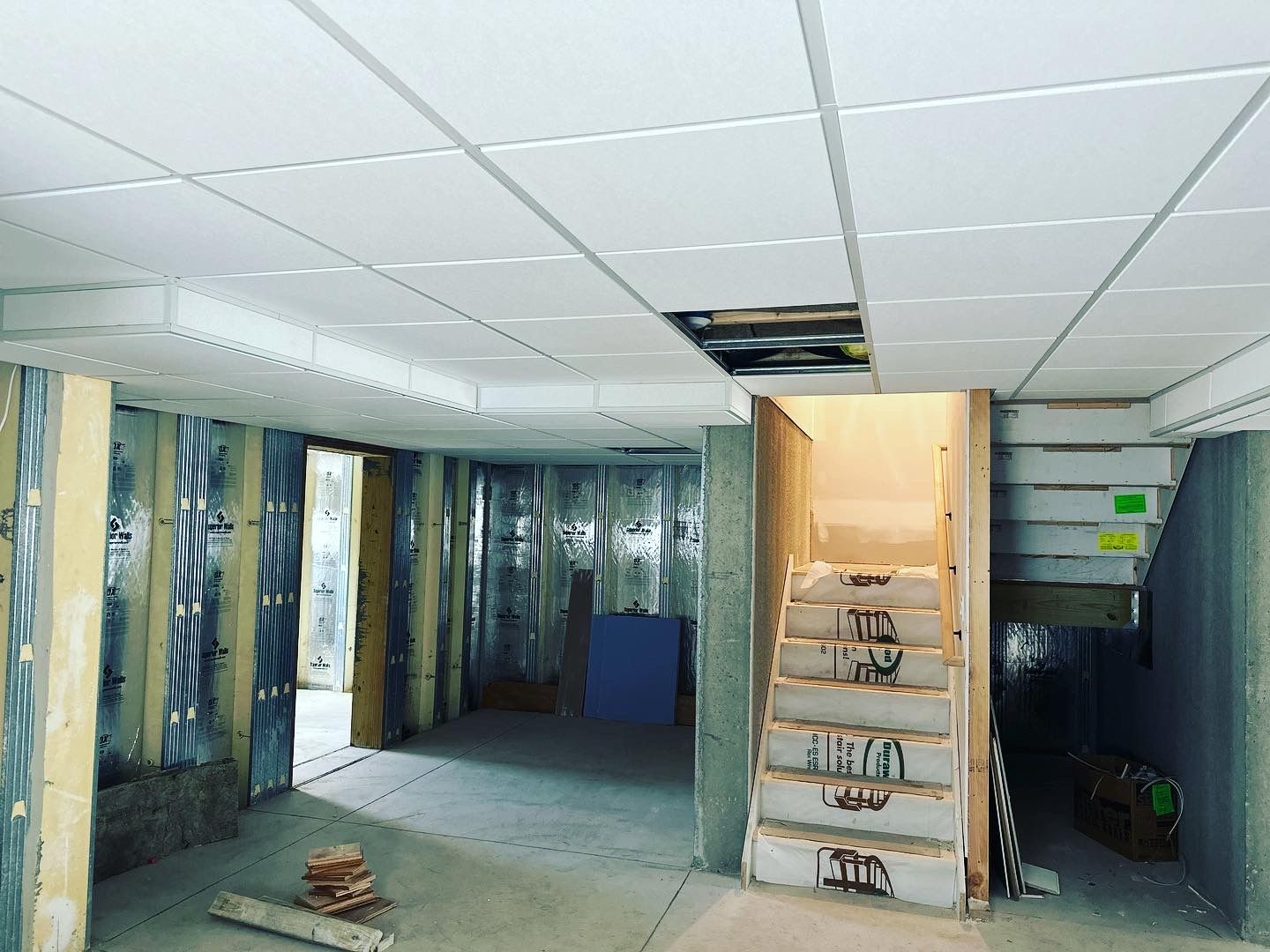 Basement under construction: exposed studs, concrete floor, stairs, and a drop ceiling.