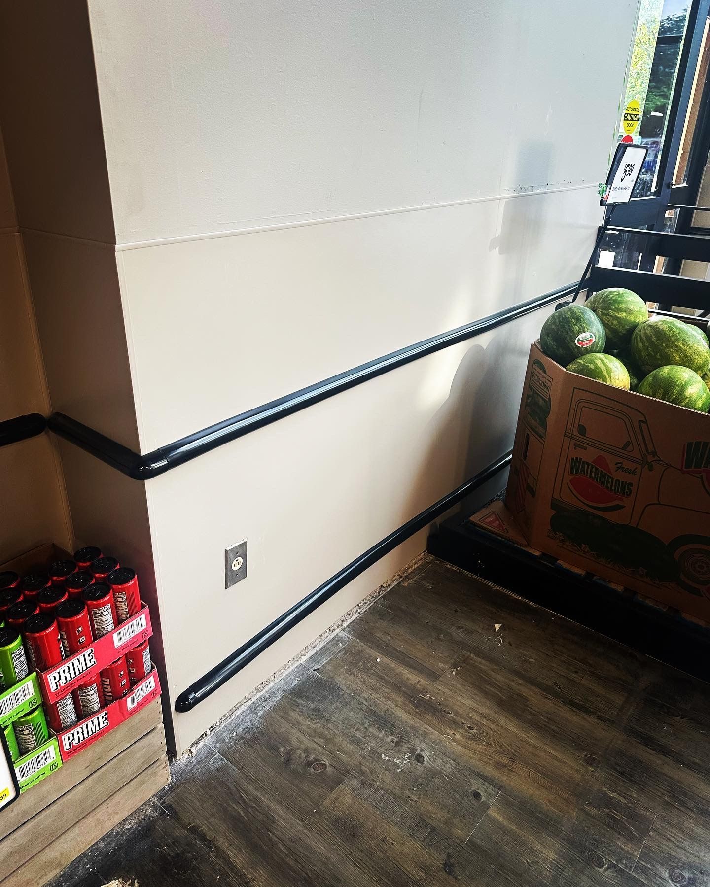 A grocery store corner with black metal bars on the wall. Watermelons and canned drinks are visible.