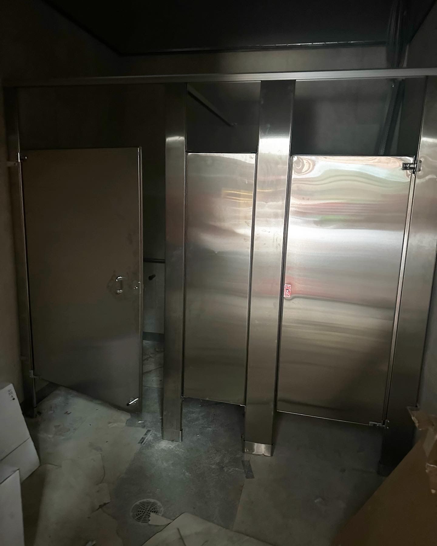 Stainless steel public restroom stalls in a room under construction; concrete floor.