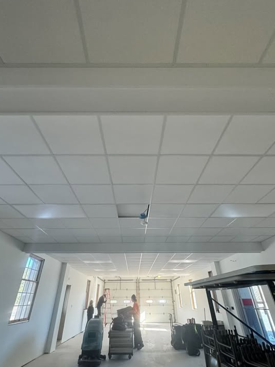 Workers in a bright room with a grid ceiling, carts, and an open garage door.